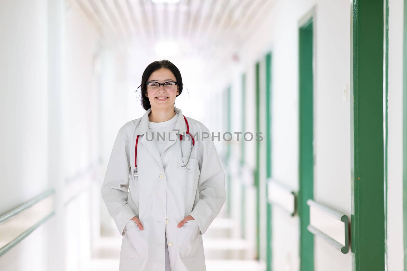Confident young female doctor with a stethoscope smiling while standing in a bright hospital corridor.