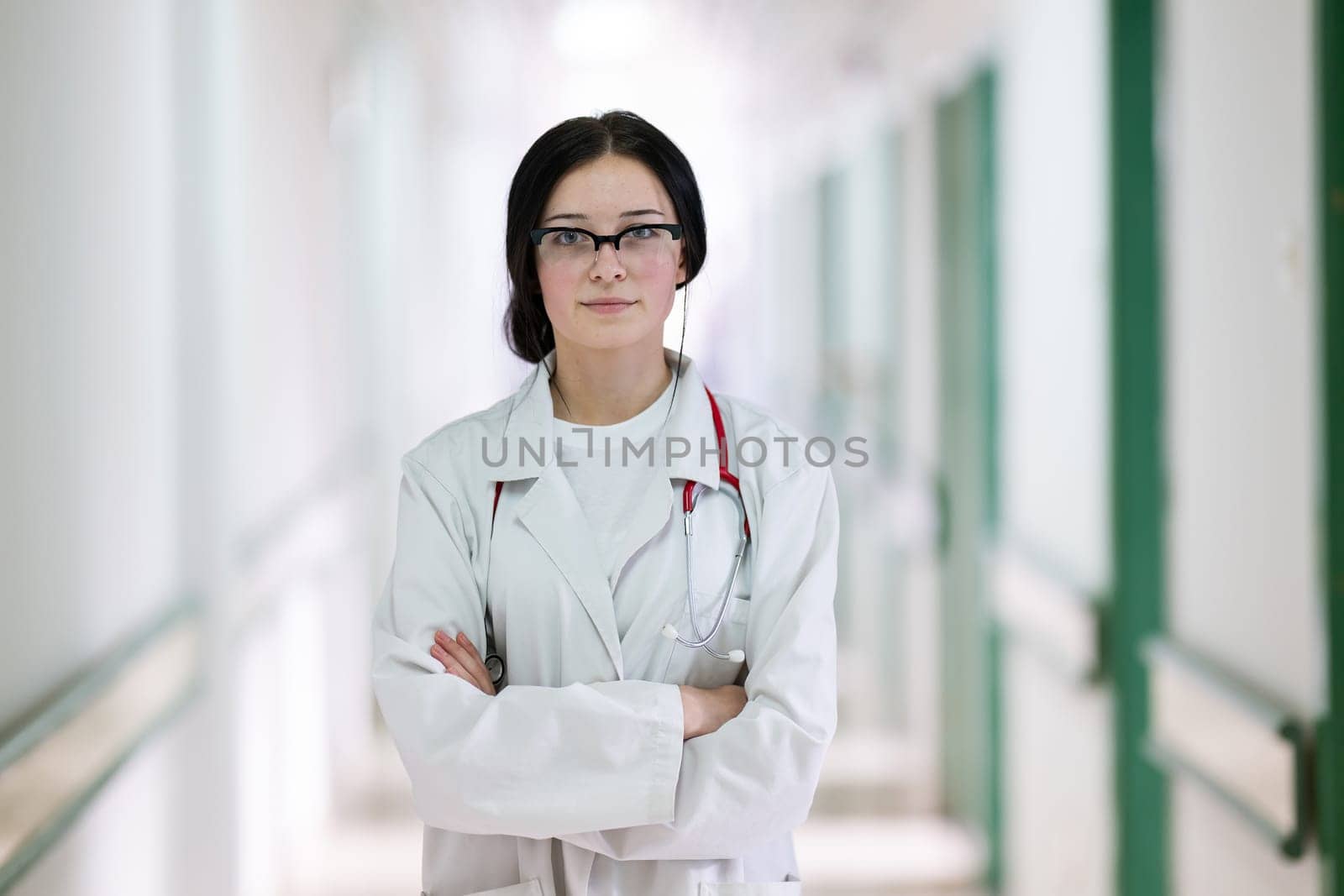 Confident young female doctor with a stethoscope smiling while standing in a bright hospital corridor.