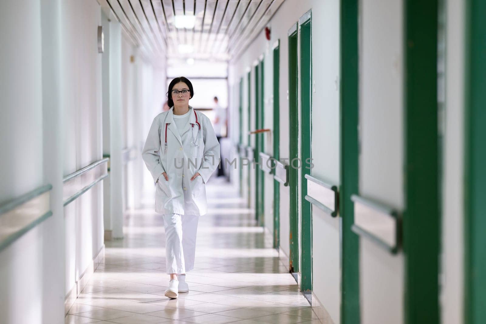 Confident young female doctor with a stethoscope smiling while standing in a bright hospital corridor.