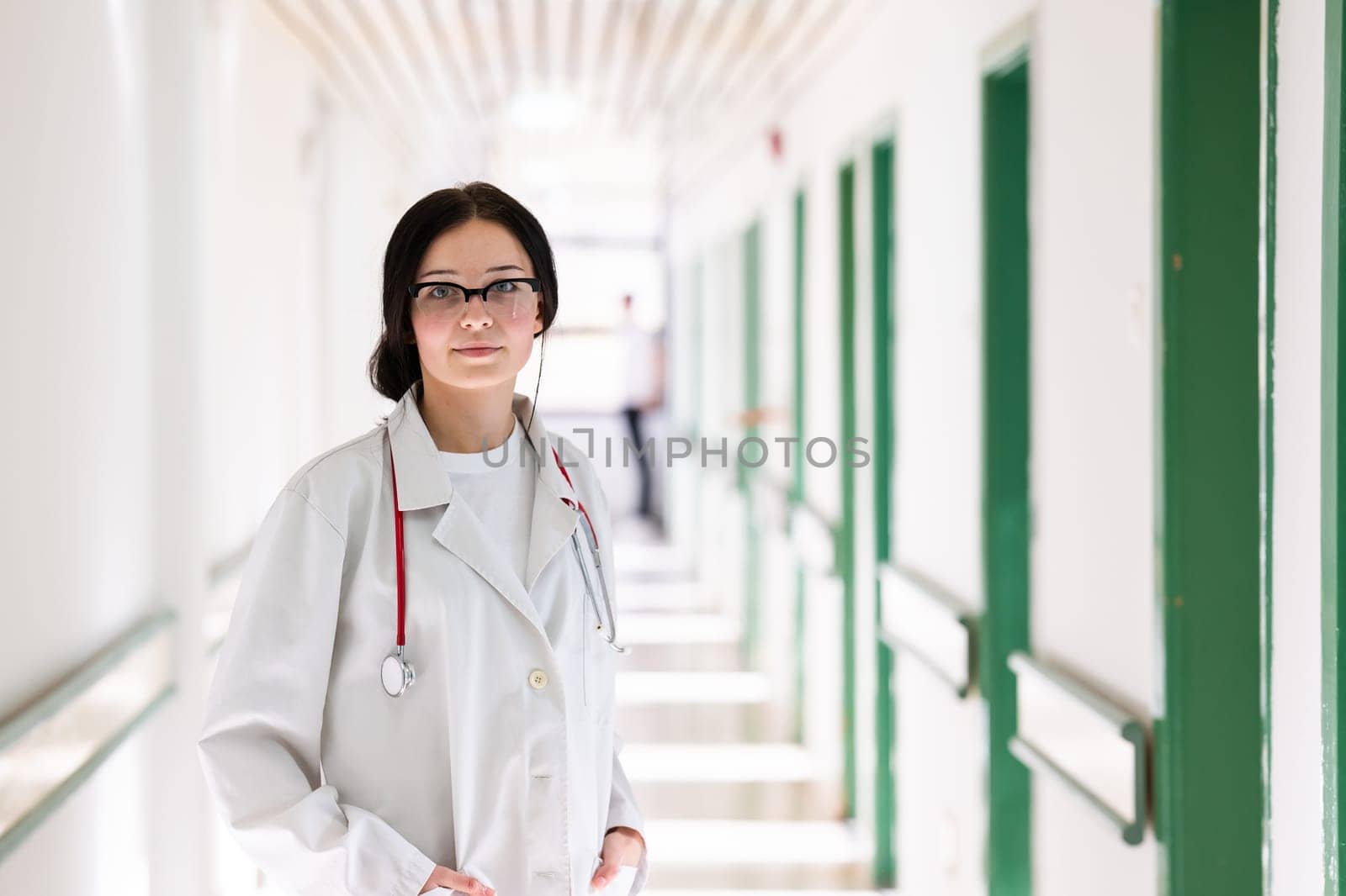 Confident young female doctor with a stethoscope smiling while standing in a bright hospital corridor.