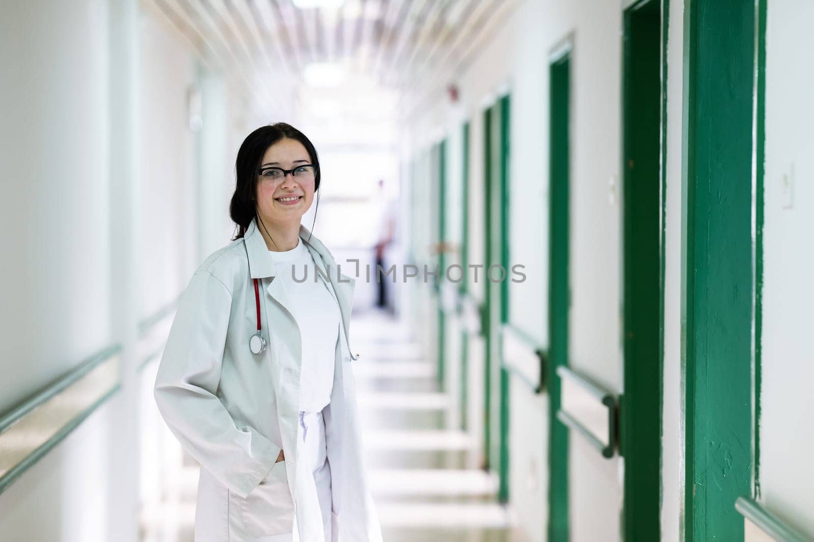 Confident young female doctor with a stethoscope smiling while standing in a bright hospital corridor.