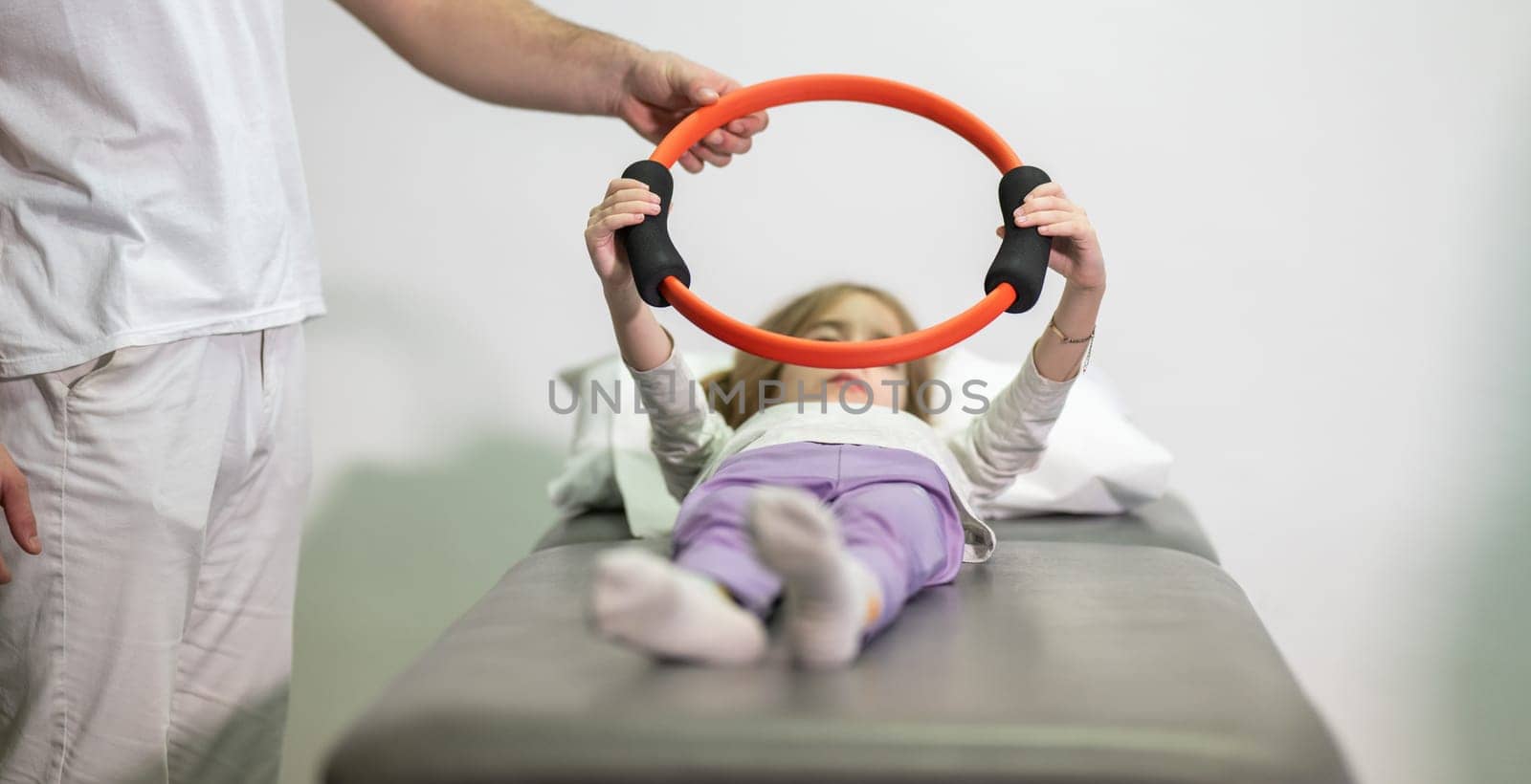 Young girl performing rehabilitation exercises with the help of a trainer, using a resistance ring while lying on a medical bench.
