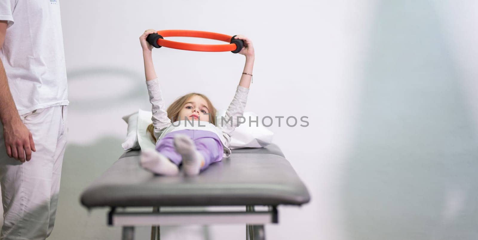 Young girl performing rehabilitation exercises with the help of a trainer, using a resistance ring while lying on a medical bench.