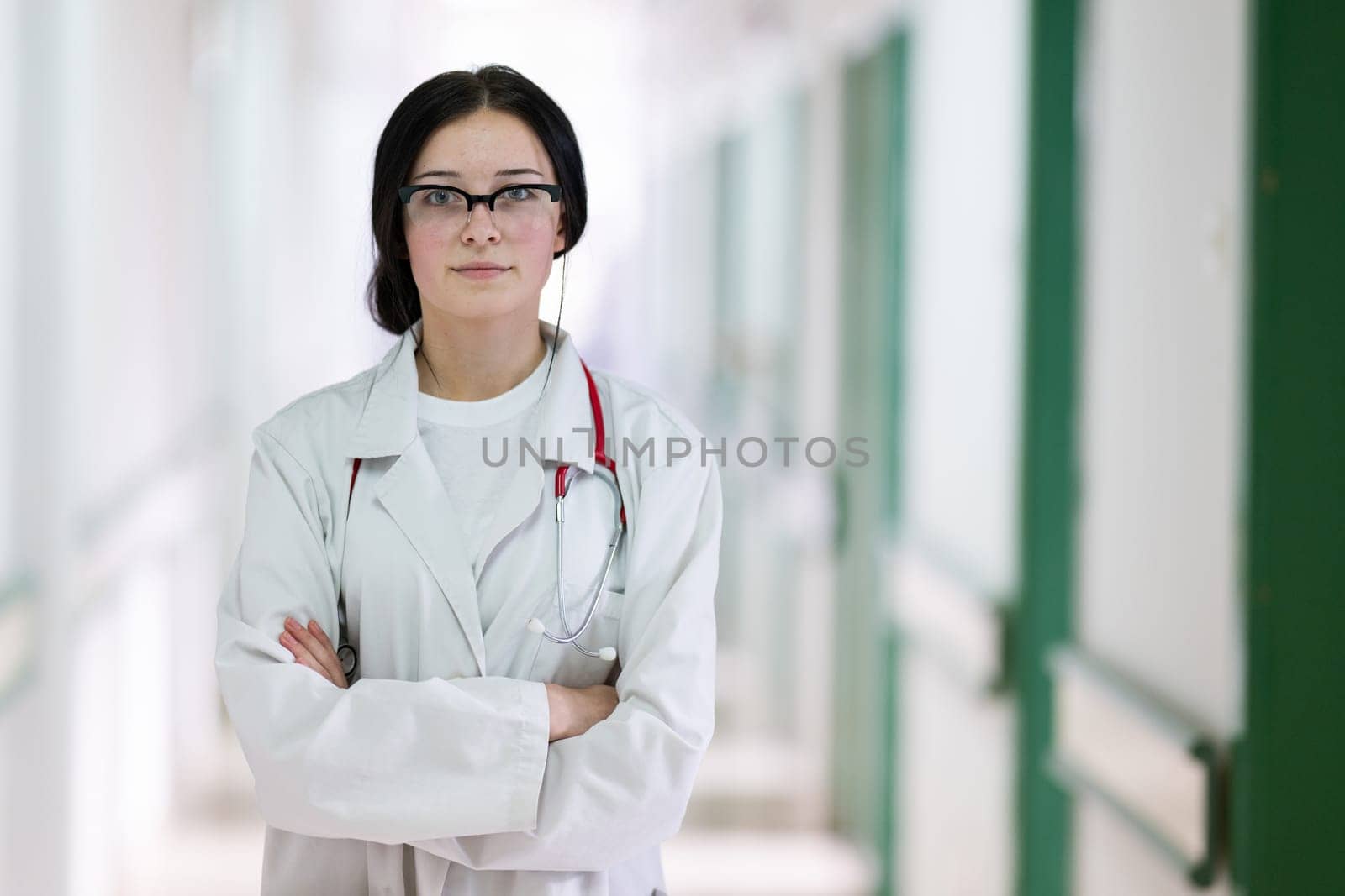 Confident young female doctor with a stethoscope smiling while standing in a bright hospital corridor.