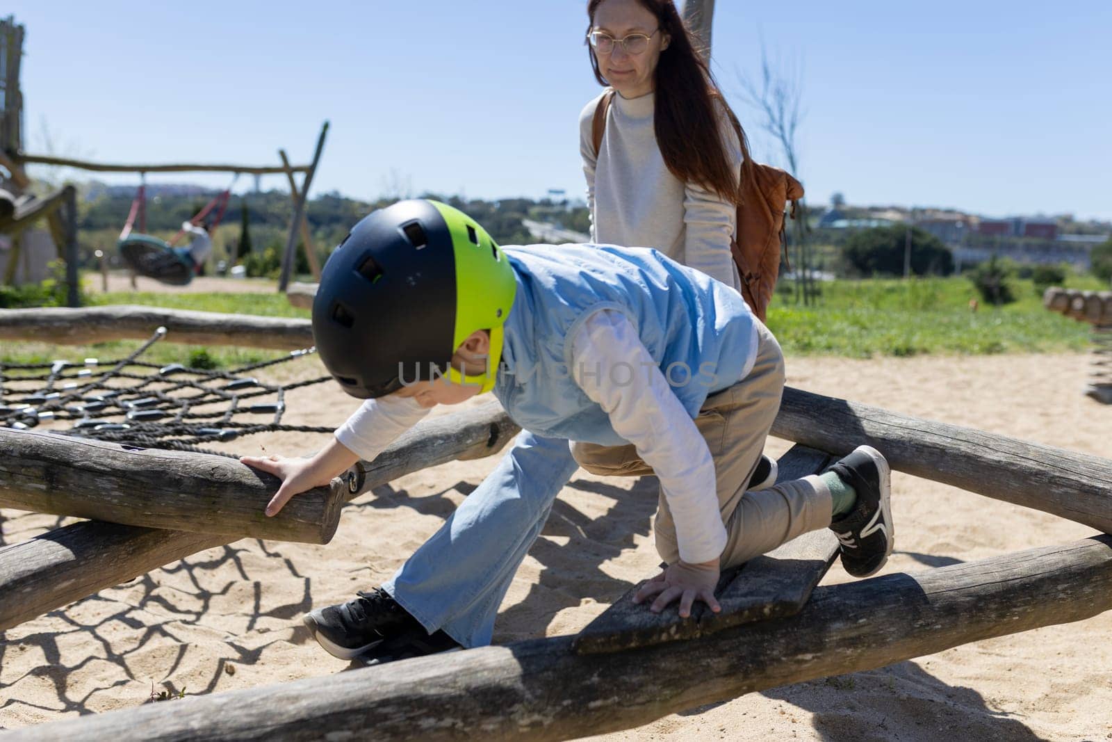 Child wearing a helmet playing on a playground climbing frame made of logs with a woman watching in the background