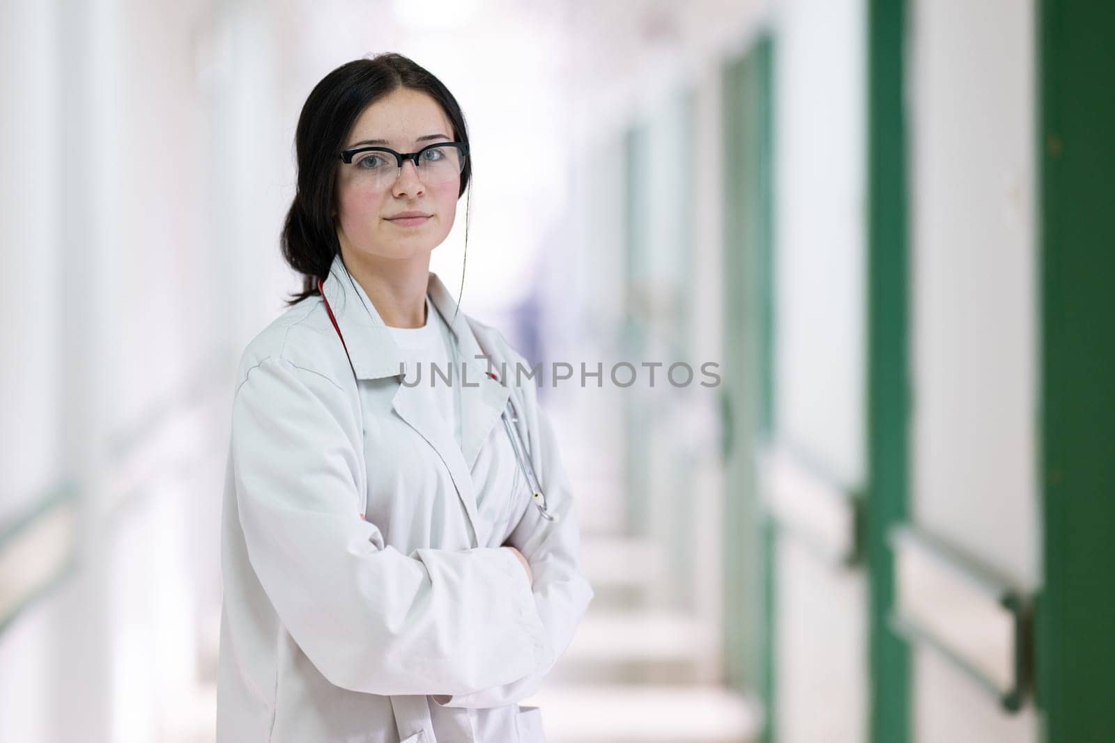 Confident young female doctor with a stethoscope smiling while standing in a bright hospital corridor.