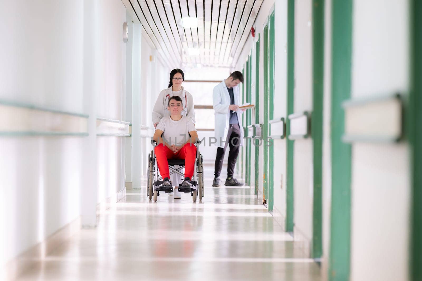 A smiling young man in a wheelchair is being pushed by a female healthcare worker in a white coat and stethoscope. They are in a hospital corridor, suggesting a medical or rehabilitation environment, evoking care and recovery.