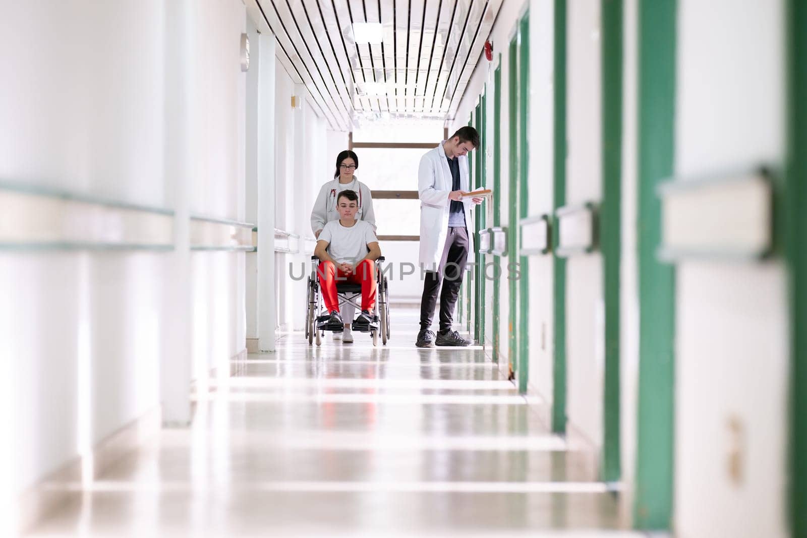 A smiling young man in a wheelchair is being pushed by a female healthcare worker in a white coat and stethoscope. They are in a hospital corridor, suggesting a medical or rehabilitation environment, evoking care and recovery.