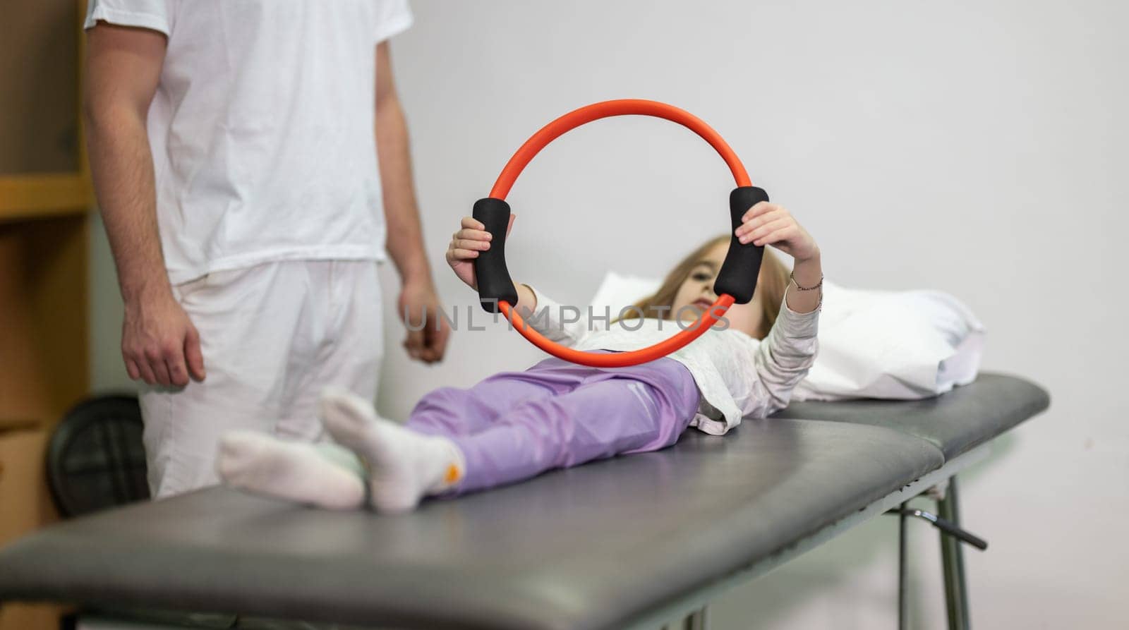 Young girl performing rehabilitation exercises with the help of a trainer, using a resistance ring while lying on a medical bench.