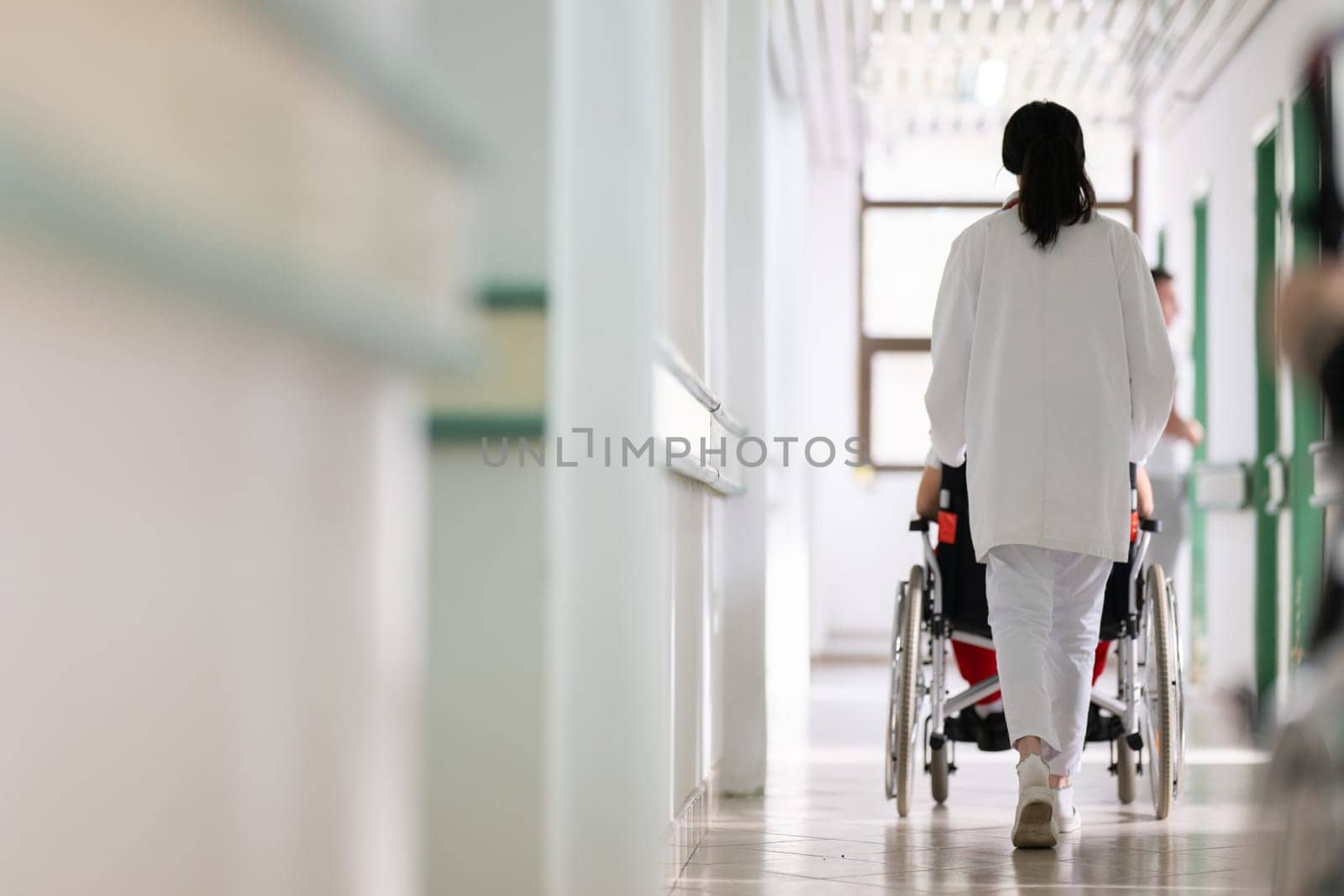A smiling young man in a wheelchair is being pushed by a female healthcare worker in a white coat and stethoscope. They are in a hospital corridor, suggesting a medical or rehabilitation environment, evoking care and recovery.