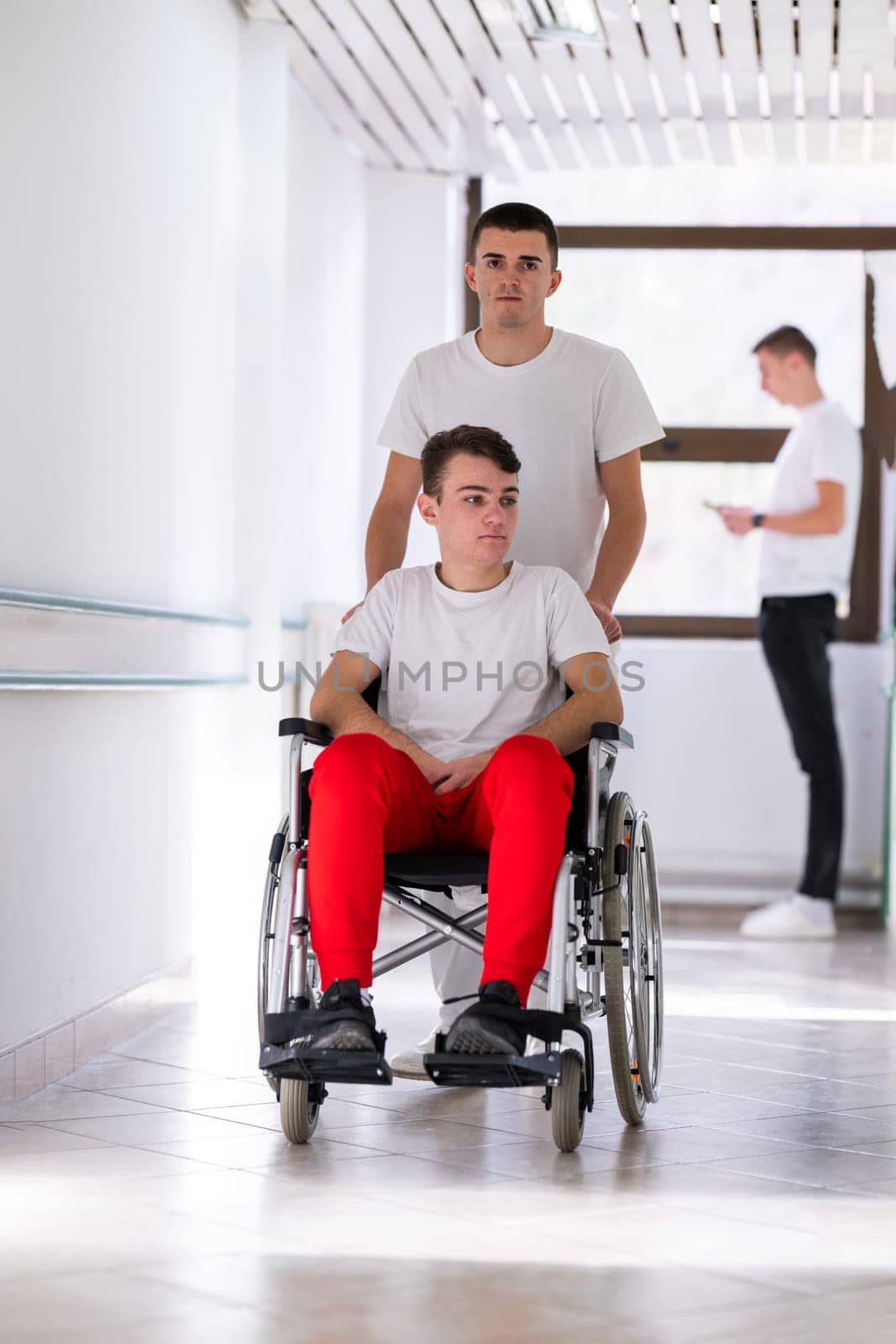 A smiling young man in a wheelchair is being pushed by a female healthcare worker in a white coat and stethoscope. They are in a hospital corridor, suggesting a medical or rehabilitation environment, evoking care and recovery.