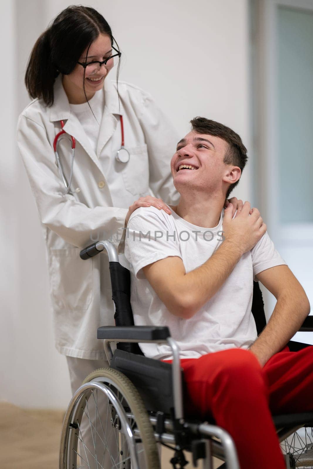 A smiling young man in a wheelchair is being pushed by a female healthcare worker in a white coat and stethoscope. They are in a hospital corridor, suggesting a medical or rehabilitation environment, evoking care and recovery.