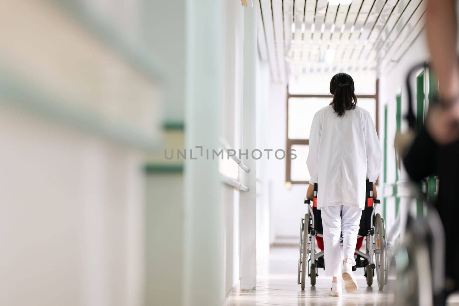 A smiling young man in a wheelchair is being pushed by a female healthcare worker in a white coat and stethoscope. They are in a hospital corridor, suggesting a medical or rehabilitation environment, evoking care and recovery.
