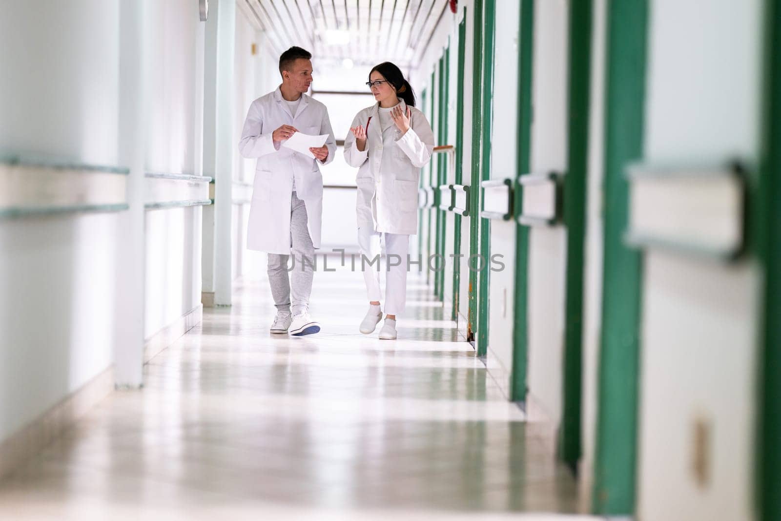 Two doctors in white coats walking and discussing medical documents in a brightly lit hospital corridor, emphasizing teamwork and professional healthcare environment.