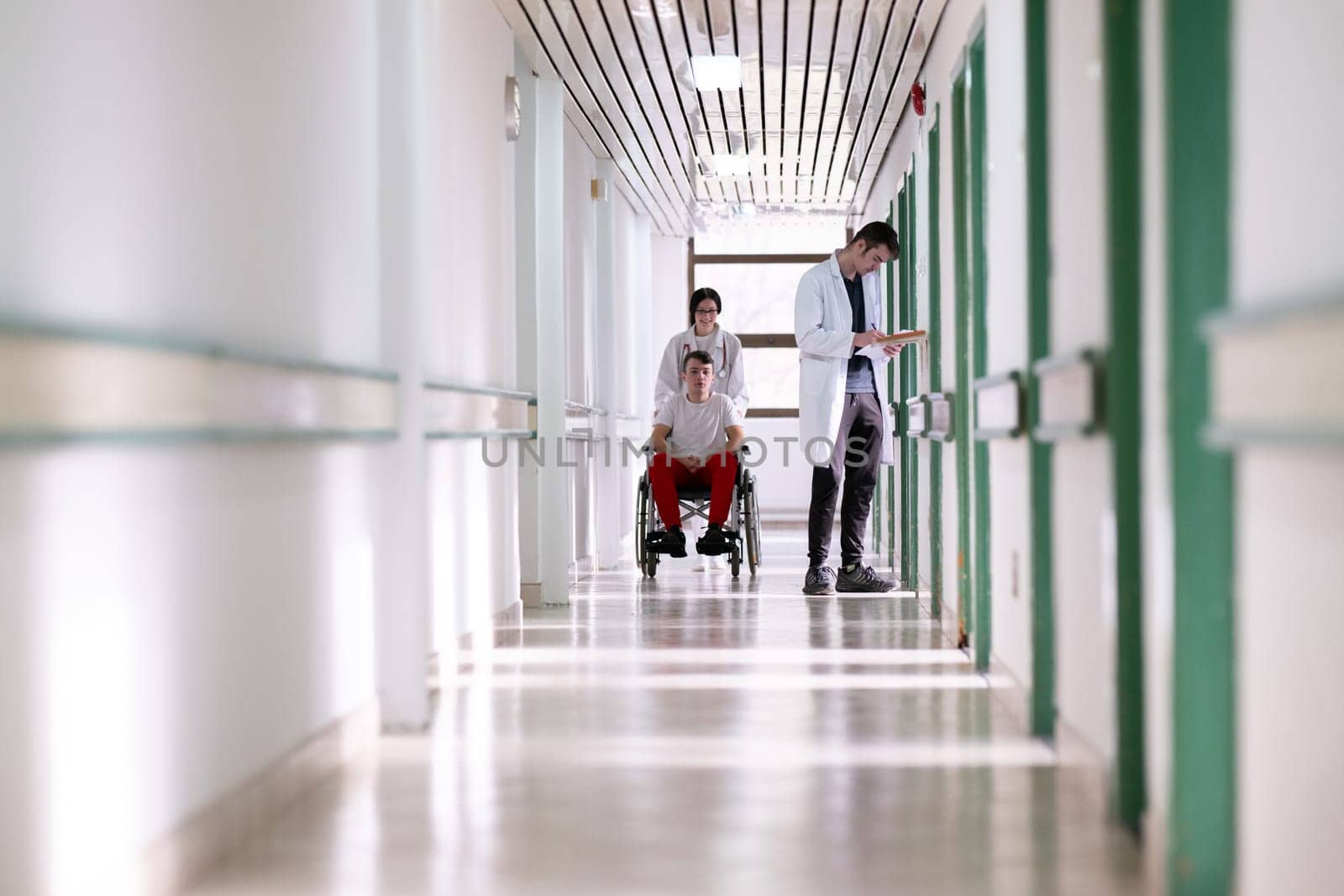 A smiling young man in a wheelchair is being pushed by a female healthcare worker in a white coat and stethoscope. They are in a hospital corridor, suggesting a medical or rehabilitation environment, evoking care and recovery.