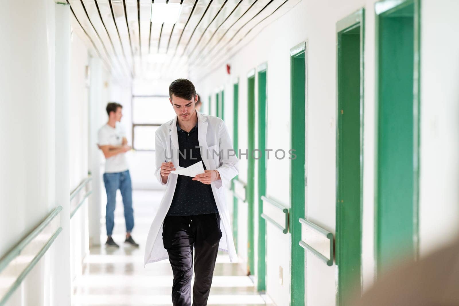 Young male doctor walking in hospital hallway while reading patient documentation. Clinical work environment.