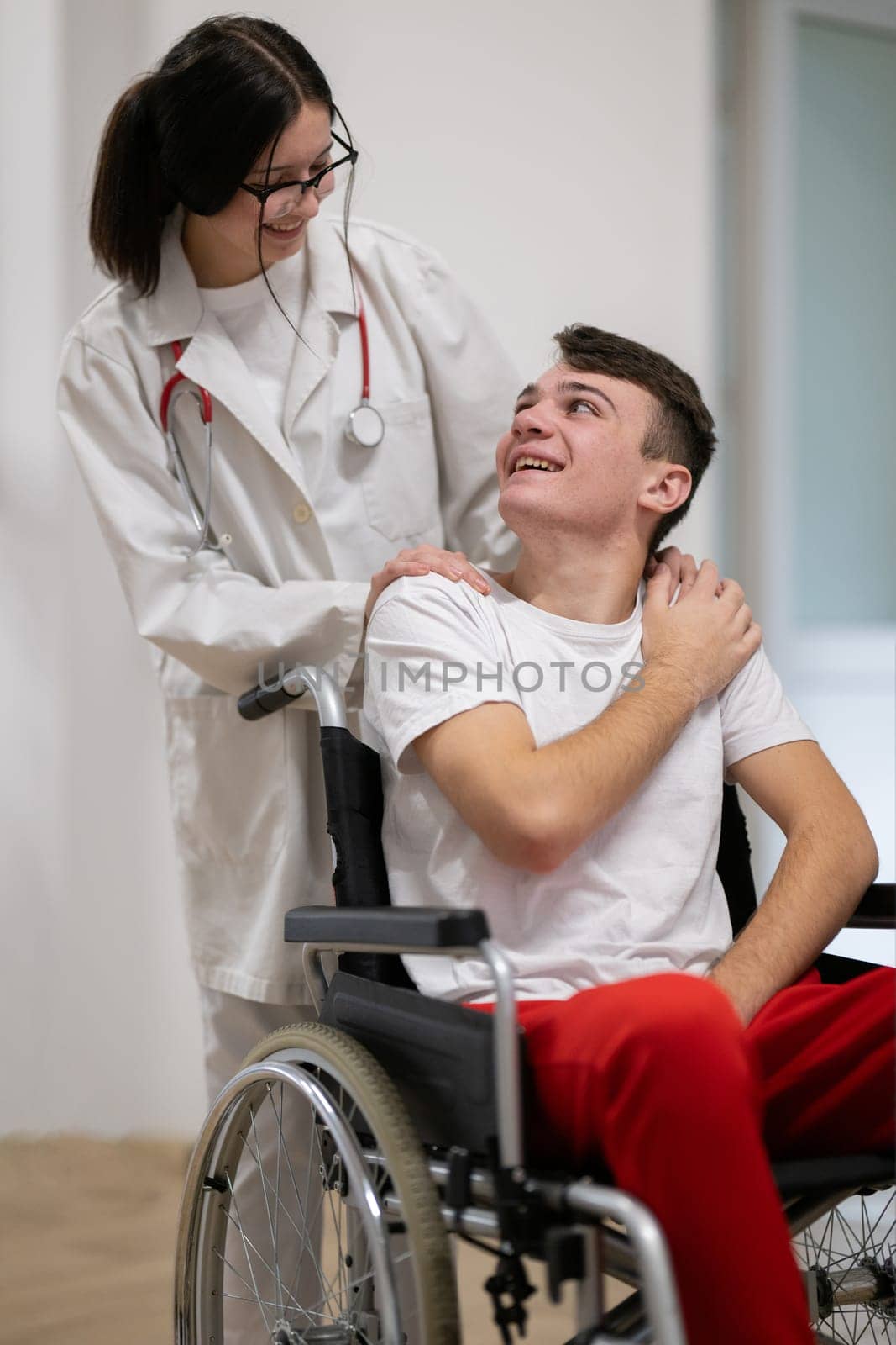 A smiling young man in a wheelchair is being pushed by a female healthcare worker in a white coat and stethoscope. They are in a hospital corridor, suggesting a medical or rehabilitation environment, evoking care and recovery.