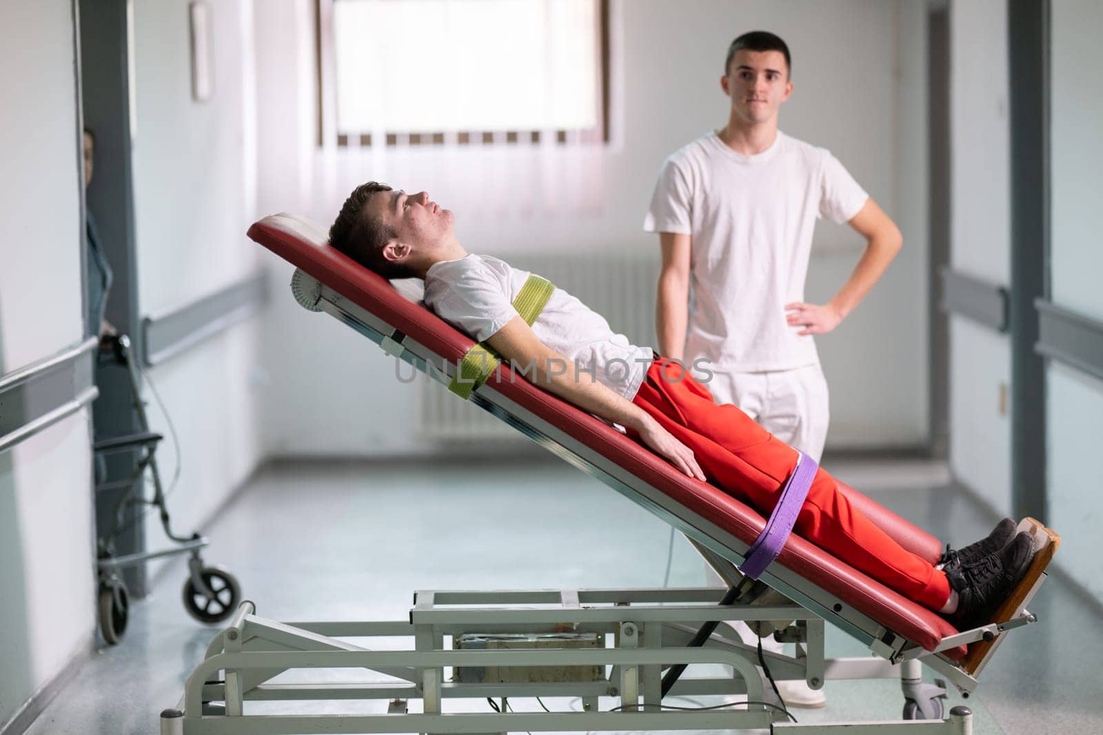 Male therapist assists patient with a standing frame during a rehabilitation session in a clinical hallway. Physical recovery support concept.