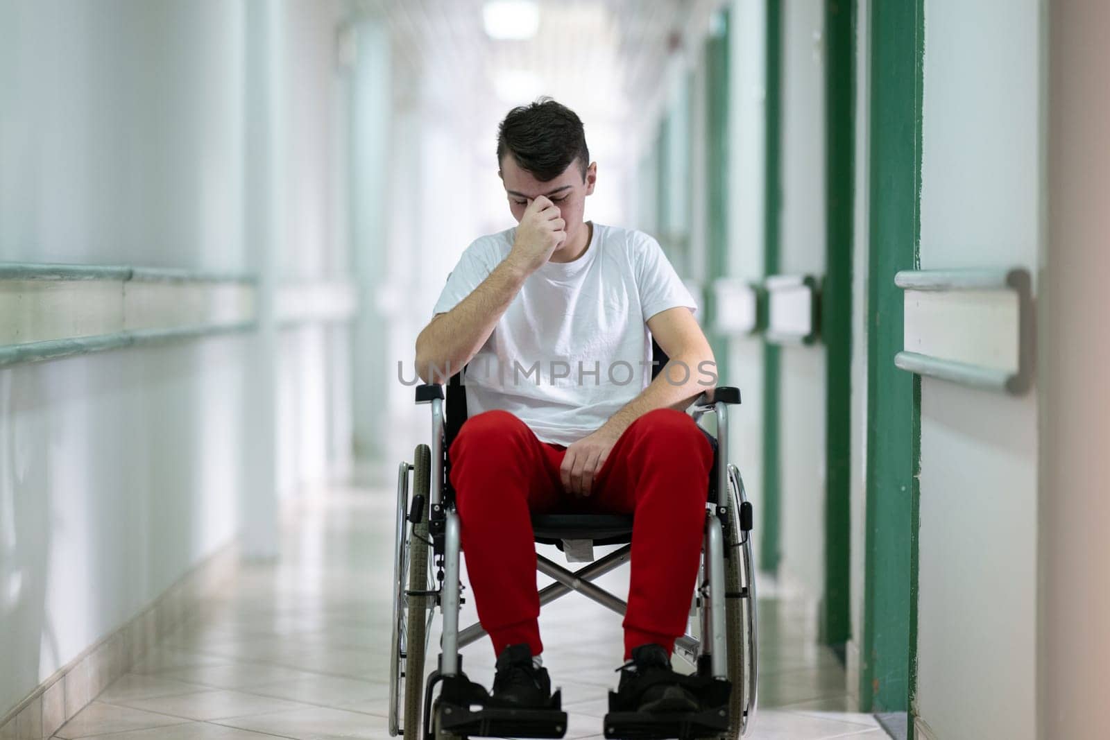 Teenage boy sitting in wheelchair with sad expression in hospital hallway. Emotion and struggle in recovery.