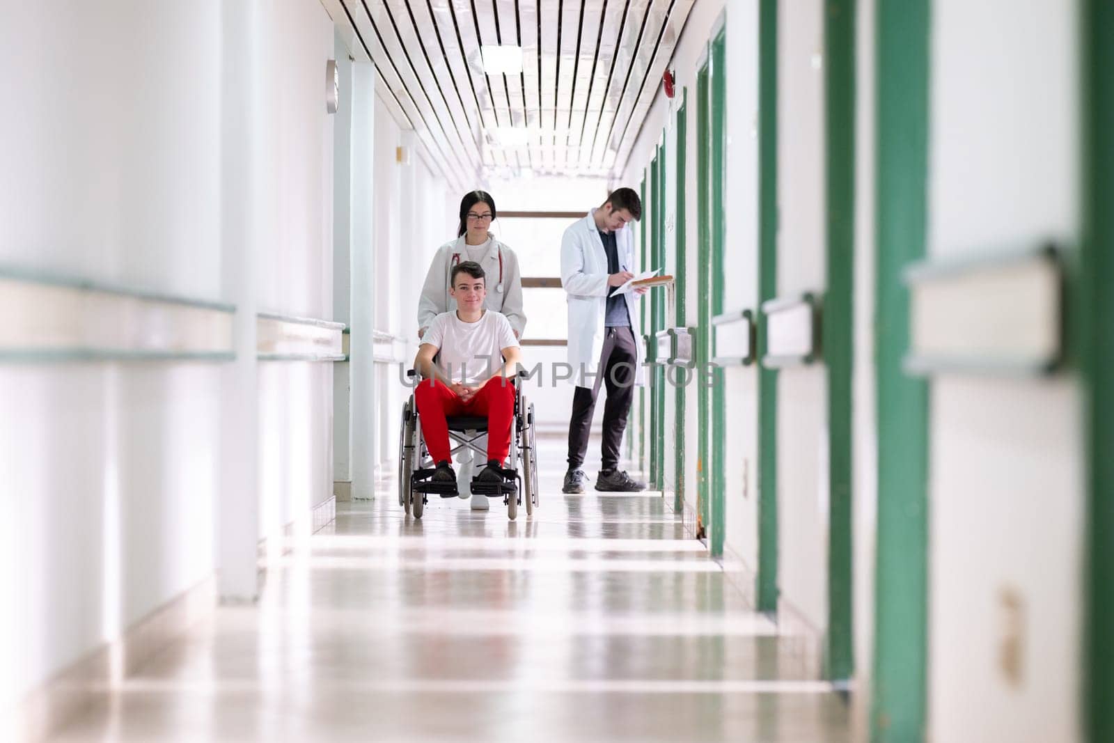 A smiling young man in a wheelchair is being pushed by a female healthcare worker in a white coat and stethoscope. They are in a hospital corridor, suggesting a medical or rehabilitation environment, evoking care and recovery.