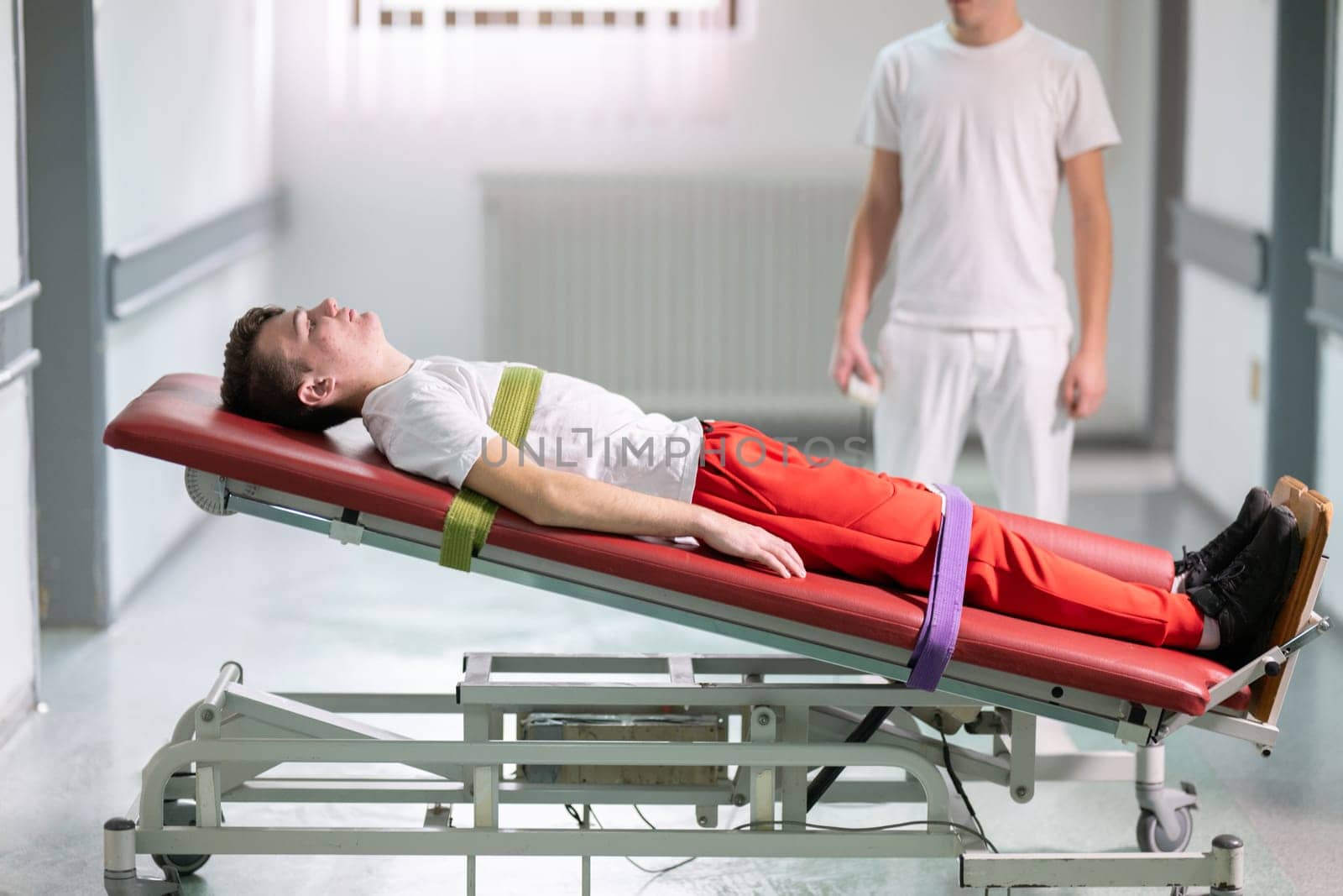 Male therapist assists patient with a standing frame during a rehabilitation session in a clinical hallway. Physical recovery support concept.