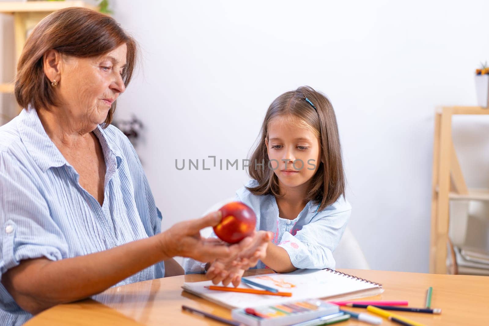 Grandmother gently places apple in girl's hand as she sits at table with colored pencils and book, creating warm, nurturing moment filled with creativity and care