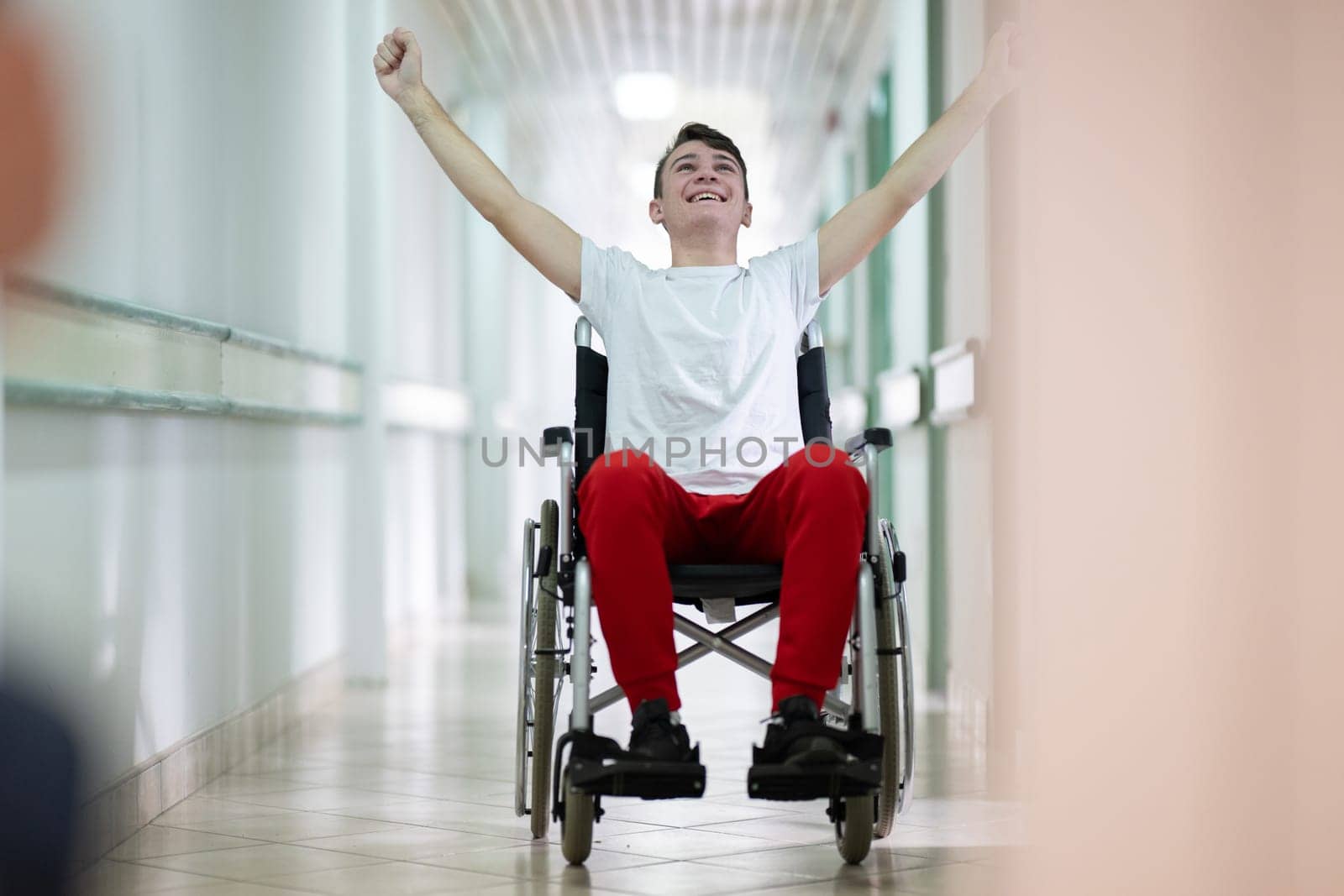 Teenage boy celebrating in wheelchair with raised arms in hospital hallway. Positivity and recovery.