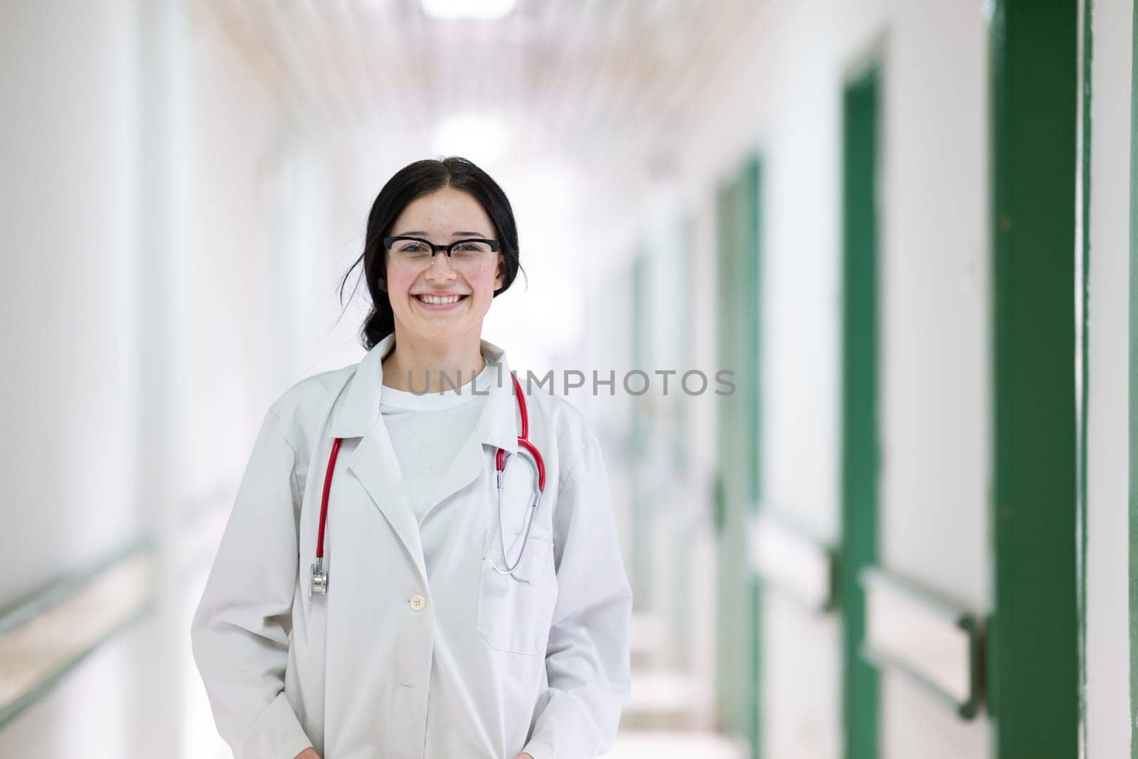 Confident young female doctor with a stethoscope smiling while standing in a bright hospital corridor.