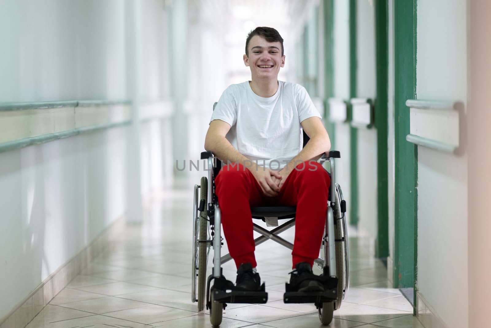 Young teenage boy smiling while sitting in a wheelchair in a hospital hallway. Rehabilitation and positivity concept