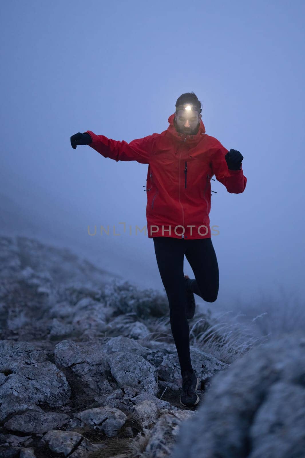 A lone hiker balances on a rocky mountain trail during early morning or dusk, with a headlamp lighting the misty path.