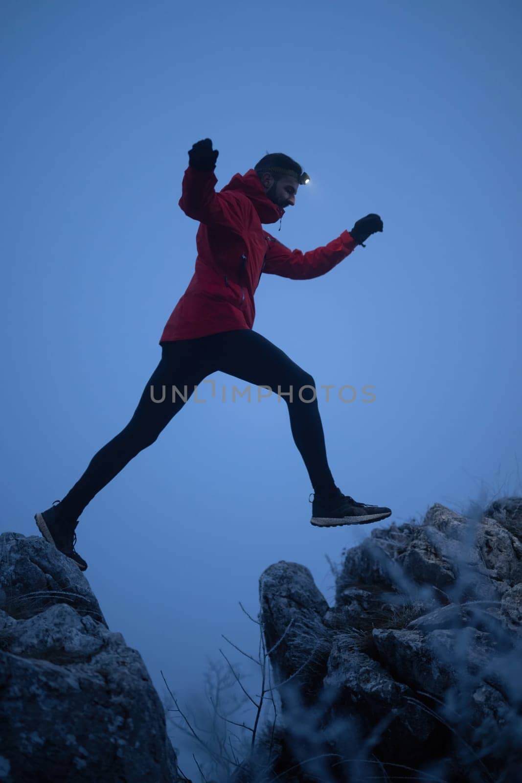 A lone hiker balances on a rocky mountain trail during early morning or dusk, with a headlamp lighting the misty path.