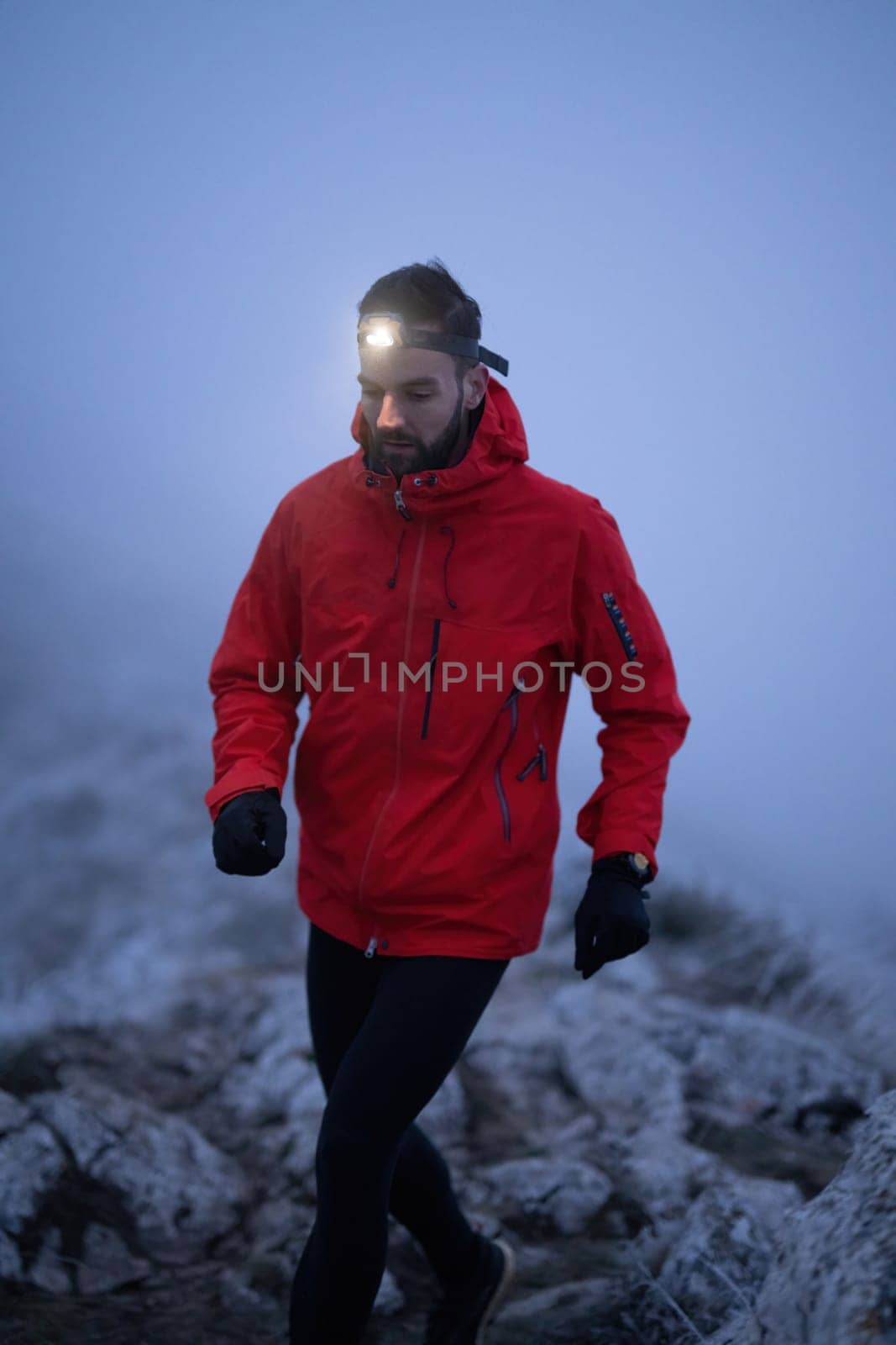 A lone hiker balances on a rocky mountain trail during early morning or dusk, with a headlamp lighting the misty path.