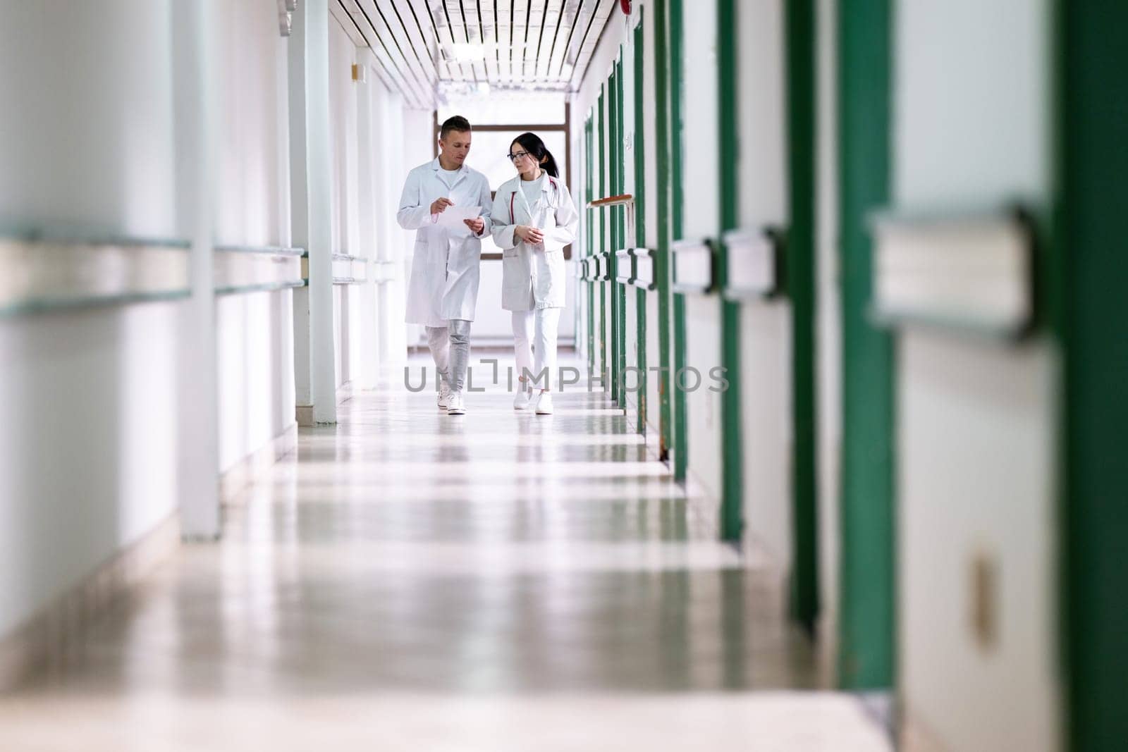 Two doctors in white coats walking and discussing medical documents in a brightly lit hospital corridor, emphasizing teamwork and professional healthcare environment.
