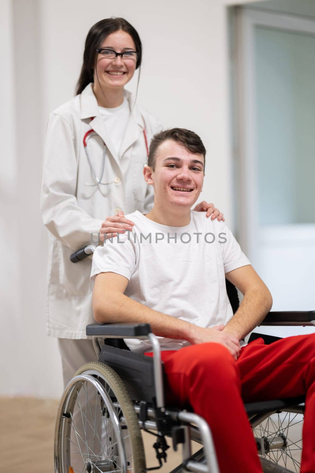 A smiling young man in a wheelchair is being pushed by a female healthcare worker in a white coat and stethoscope. They are in a hospital corridor, suggesting a medical or rehabilitation environment, evoking care and recovery.