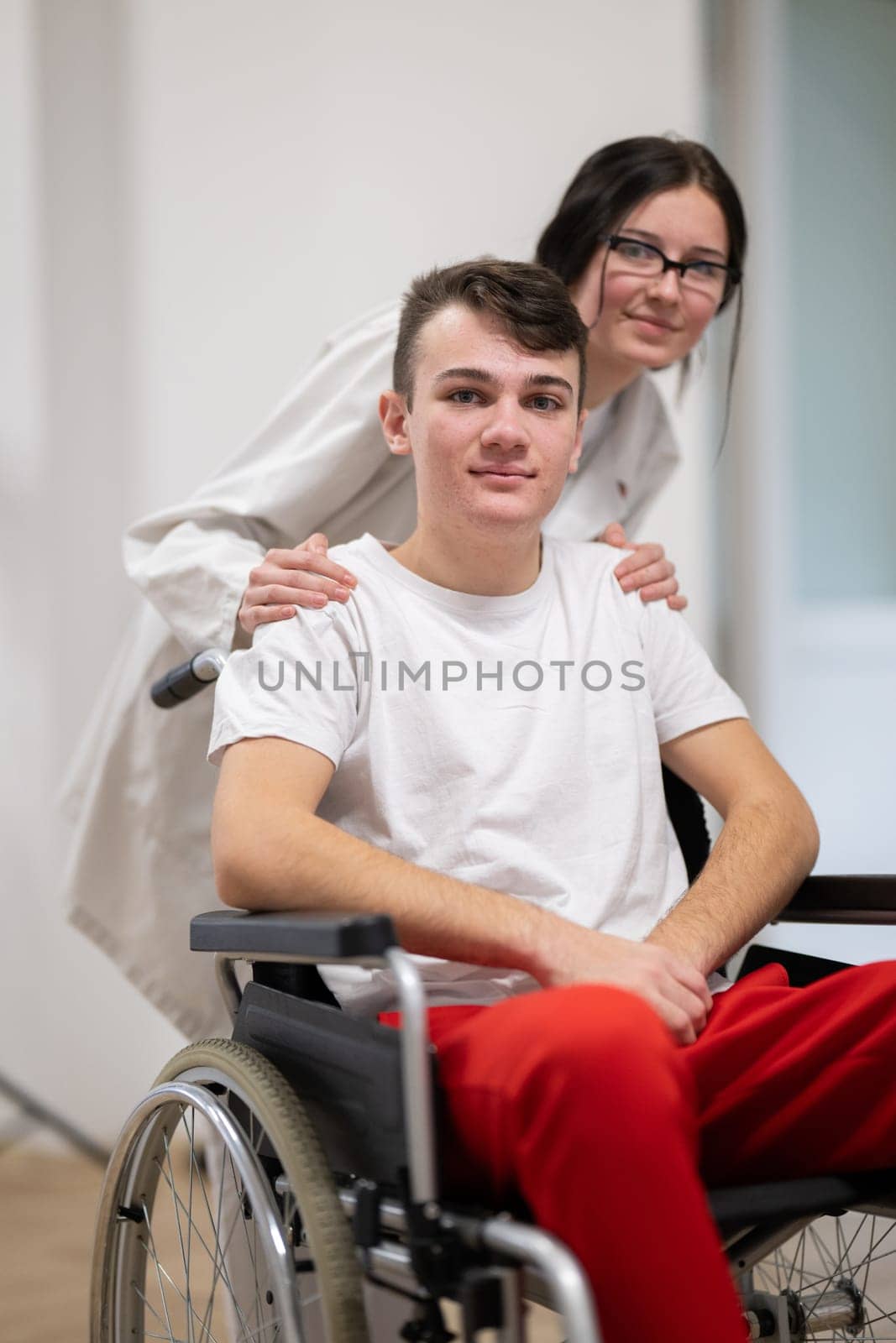 A smiling young man in a wheelchair is being pushed by a female healthcare worker in a white coat and stethoscope. They are in a hospital corridor, suggesting a medical or rehabilitation environment, evoking care and recovery.