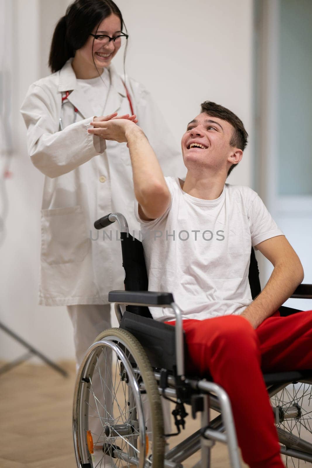 A smiling young man in a wheelchair is being pushed by a female healthcare worker in a white coat and stethoscope. They are in a hospital corridor, suggesting a medical or rehabilitation environment, evoking care and recovery.