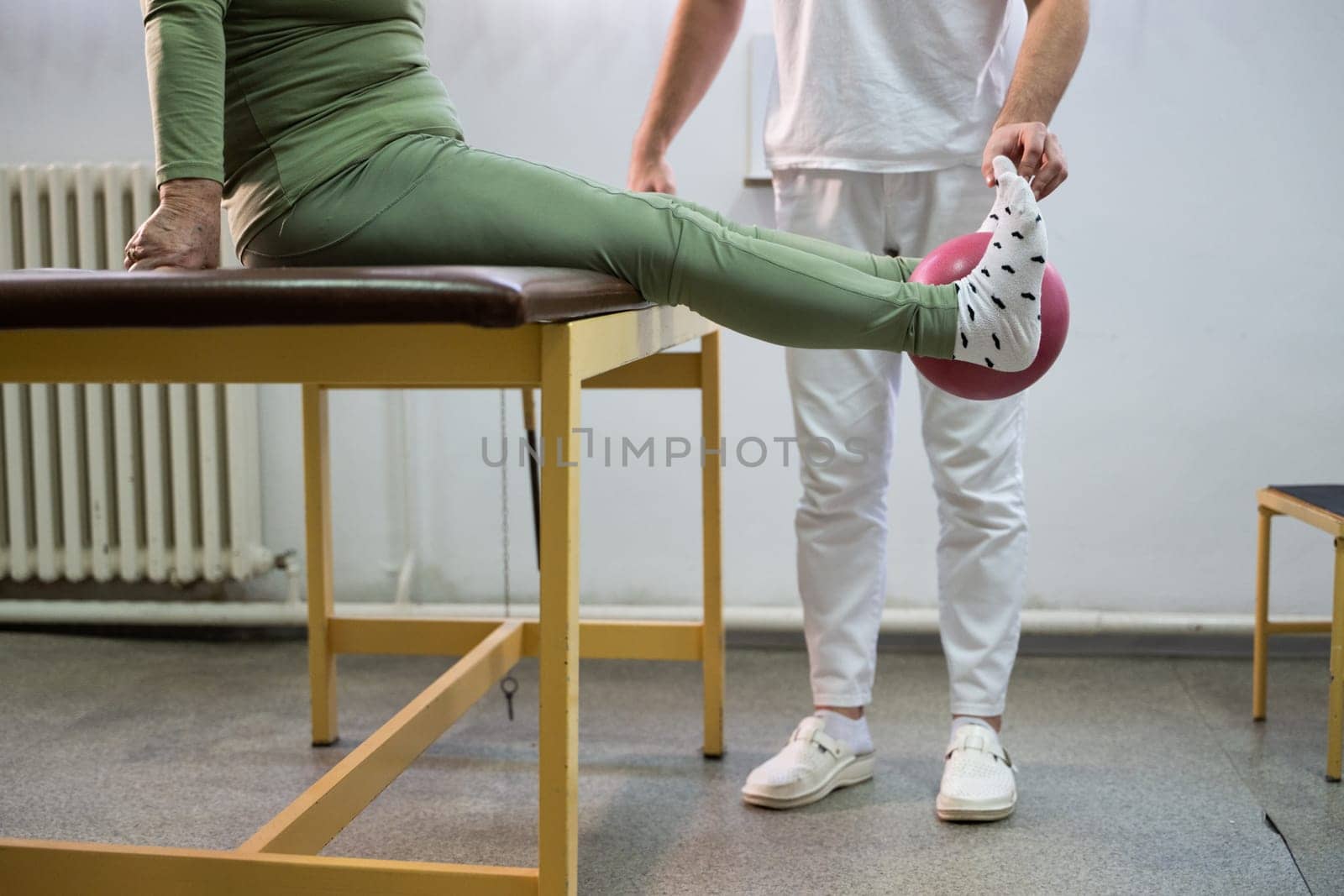 Close up of a patient performing a physical therapy exercise using a ball between the legs, assisted by a therapist.