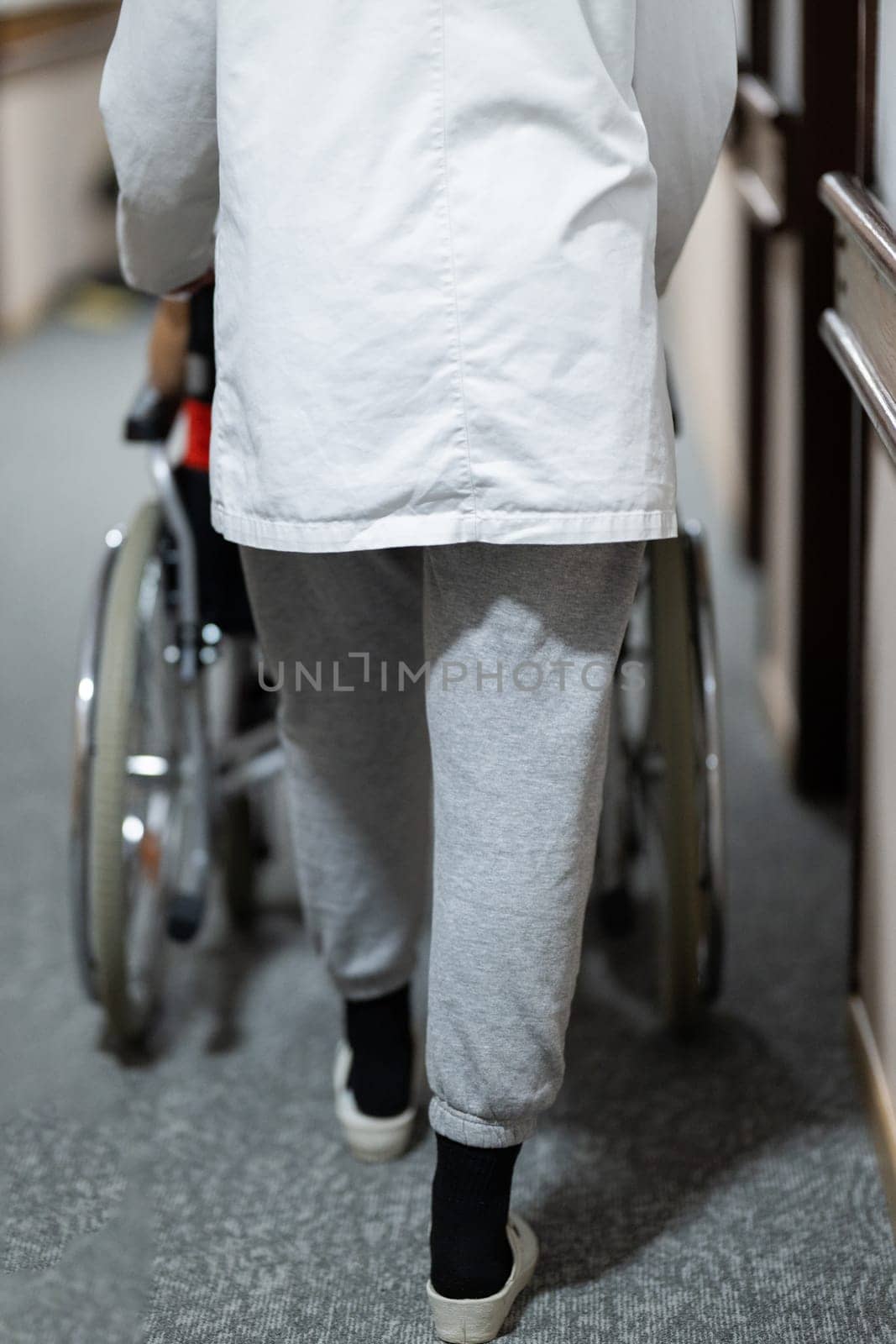A smiling young man in a wheelchair is being pushed by a female healthcare worker in a white coat and stethoscope. They are in a hospital corridor, suggesting a medical or rehabilitation environment, evoking care and recovery.