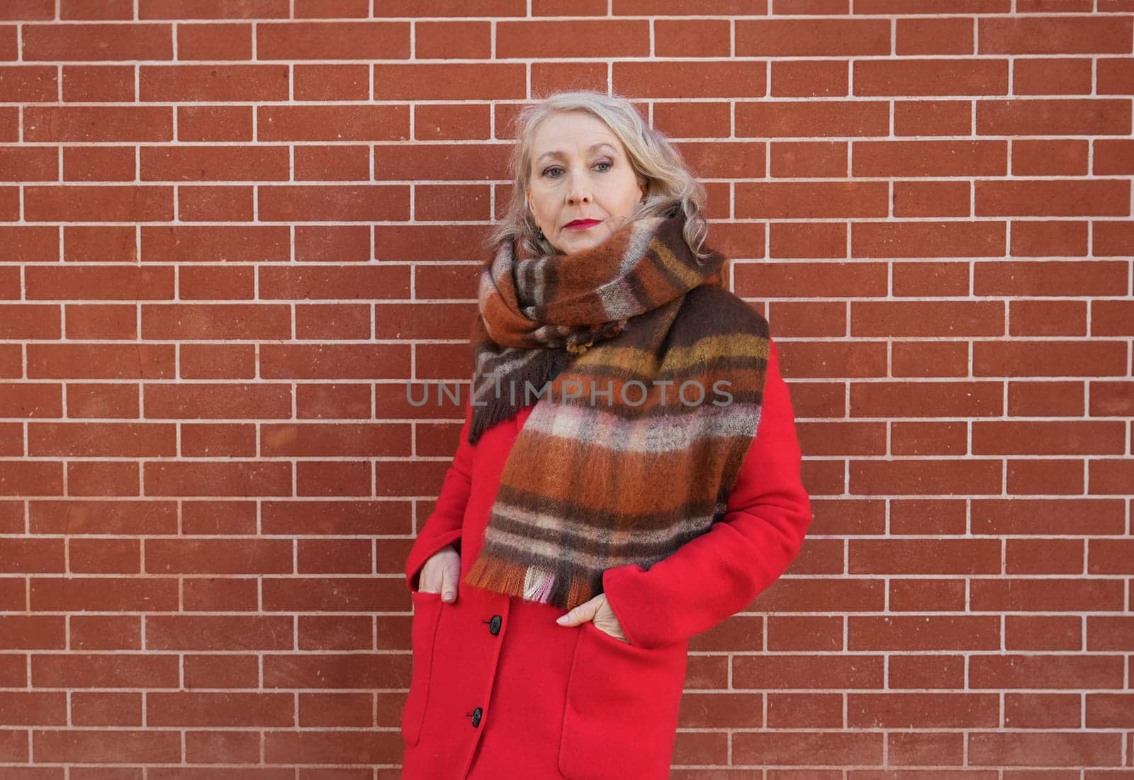 Gray-haired woman of sixty years old in a red coat and a terracotta scarf near a red -orange brick wall
