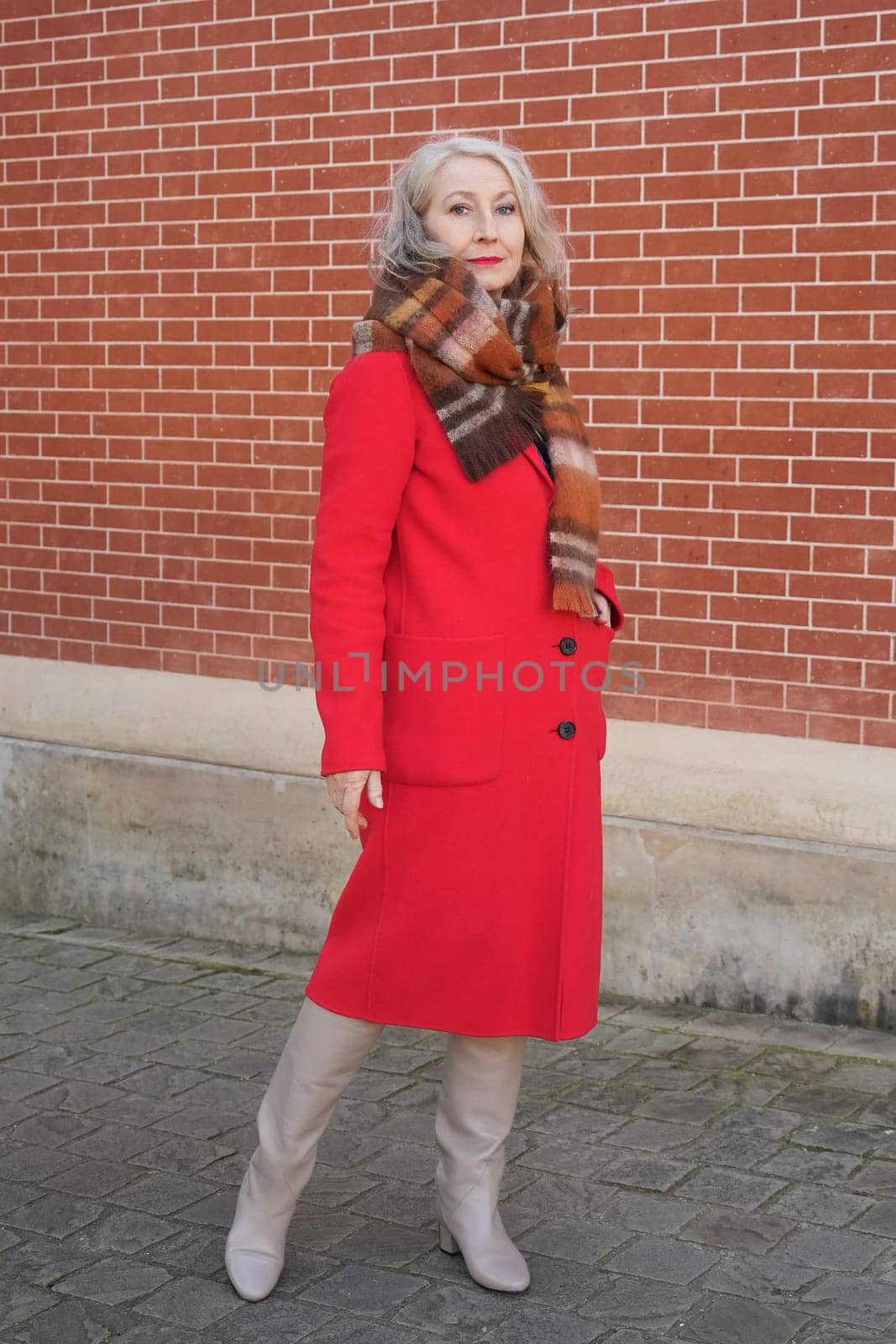 Gray-haired woman of sixty years old in a red coat and a terracotta scarf near a red -orange brick wall