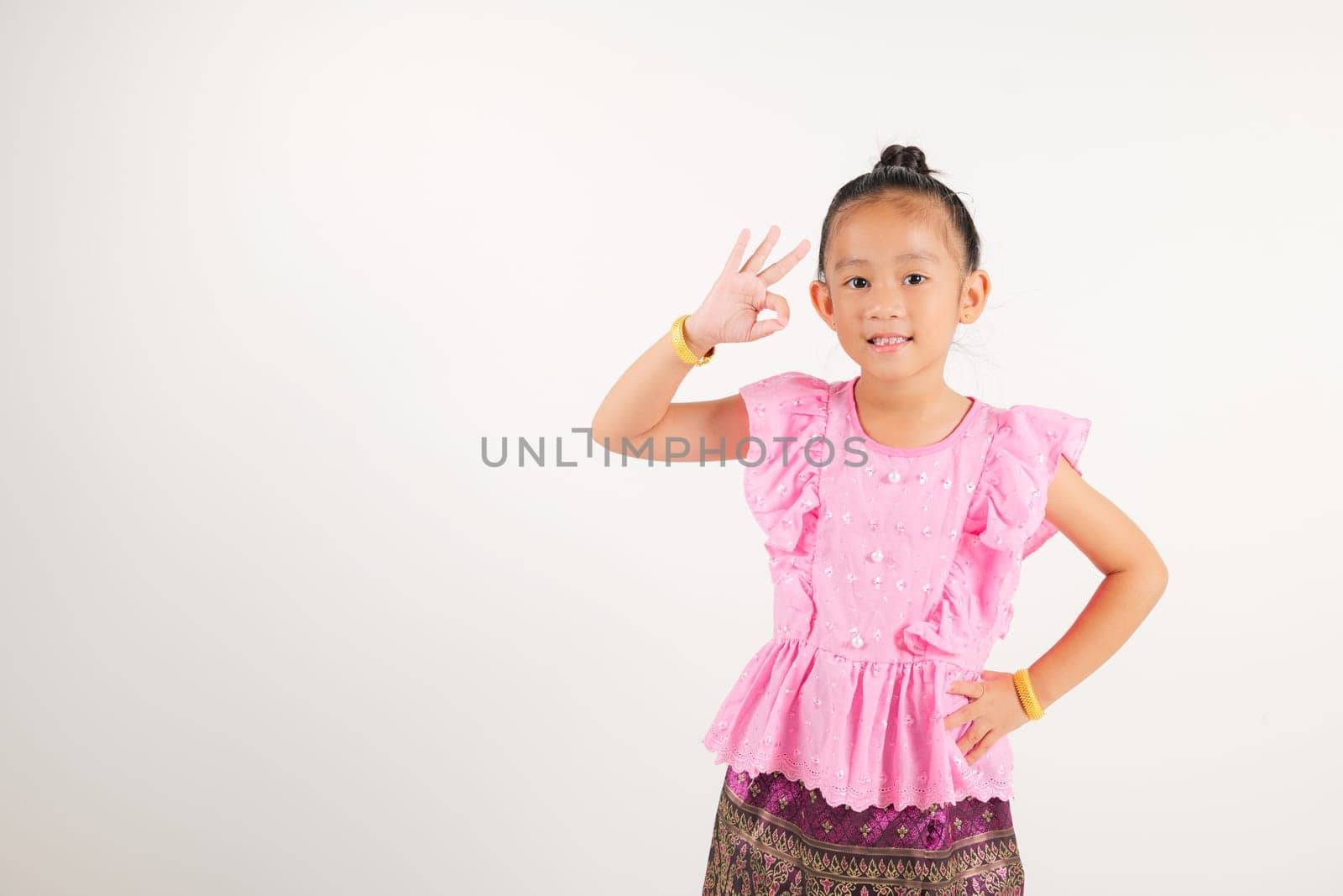 Portrait Thailand kid girl smiling traditional Thai dress costume showcases OK sign with smile, conveying agreement and positivity, studio isolated white background, kindergarten child success sign