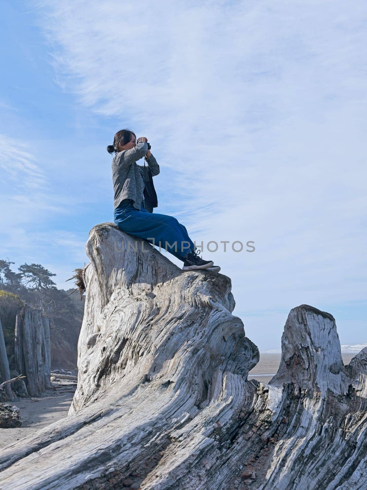 A middle aged woman looks out at the ocean with binoculars while sitting atop a large driftwood tree on a beach in Washington State.