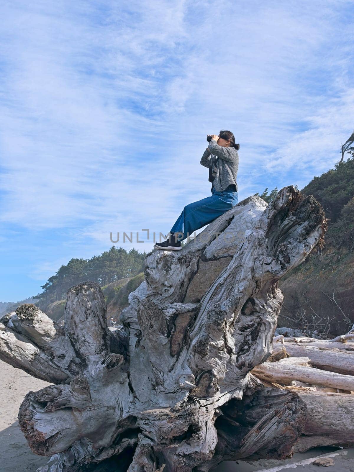 A middle aged woman looks out at the ocean with binoculars while sitting atop a large driftwood tree on a beach in Washington State.