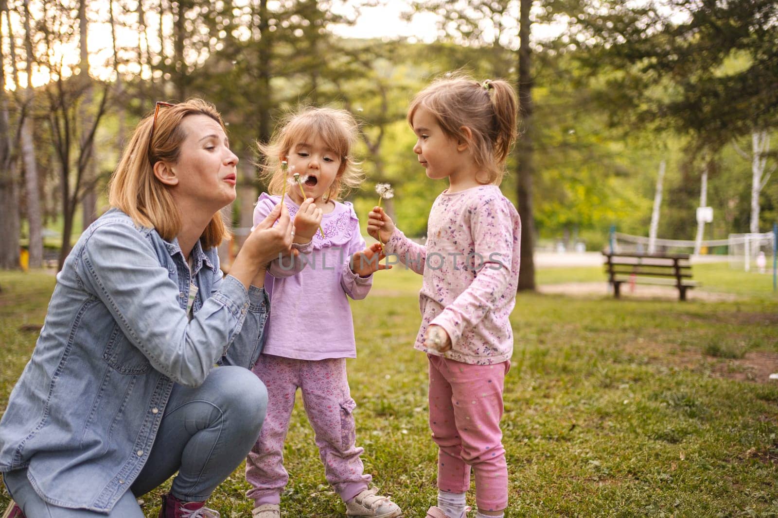 Cheerful twin daughter and mom blowing dandelions in the park.