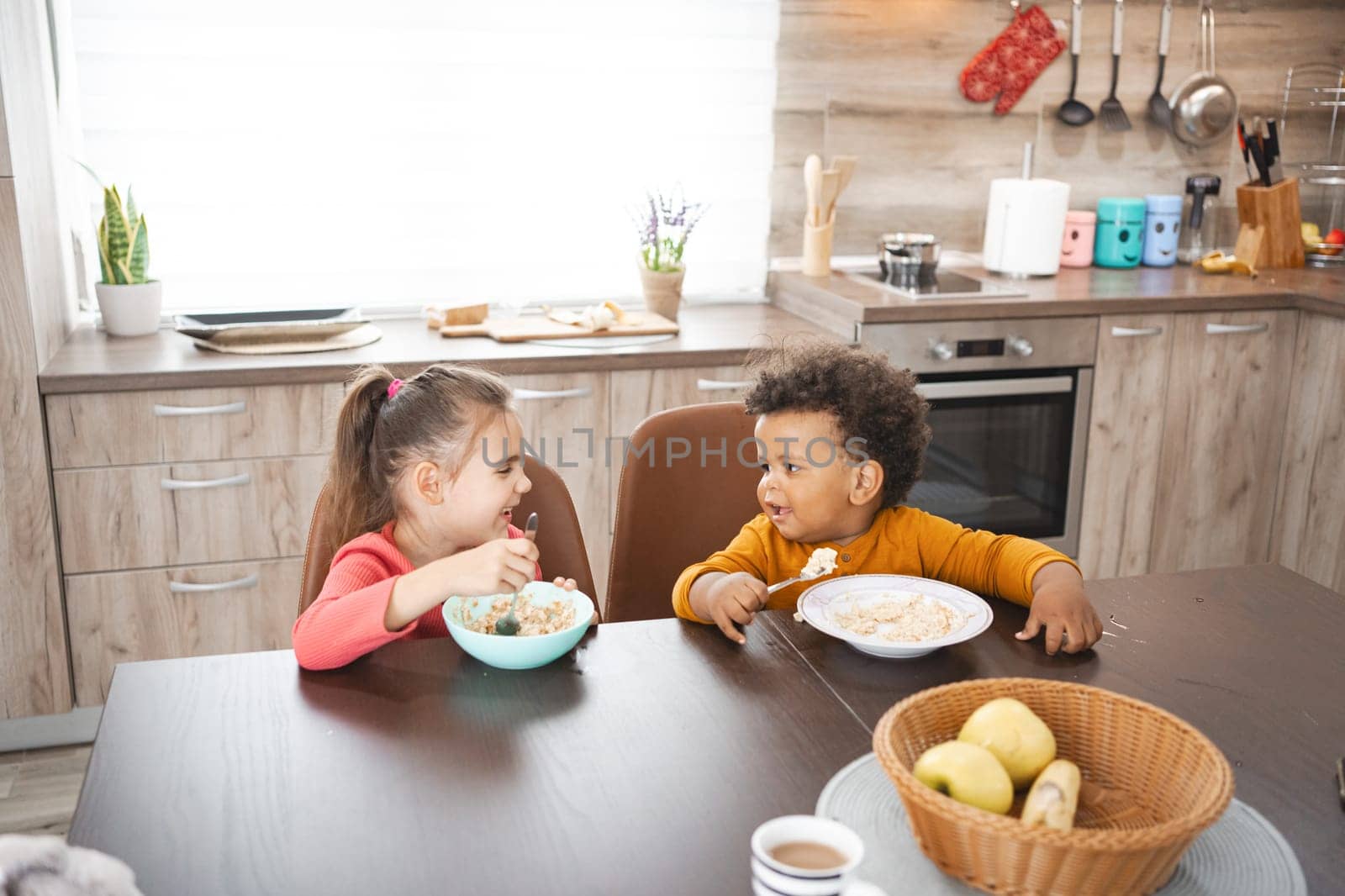 Two children share a joyful breakfast moment in a bright kitchen.