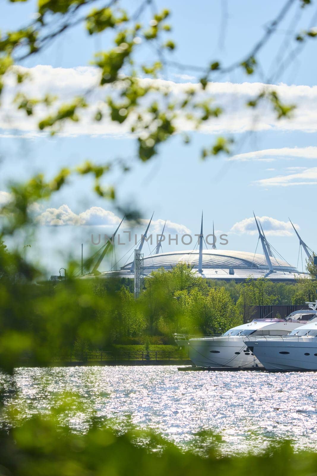 View of the mega yachts standing at the pier through the branches of trees on a clear sunny day, residential buildings and a stadium in the background, Russia, St.Petersburg. High quality photo