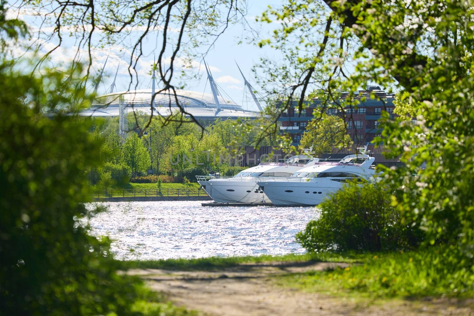 View of the mega yachts standing at the pier through the branches of trees on a clear sunny day, residential buildings and a stadium in the background, Russia, St.Petersburg. High quality photo
