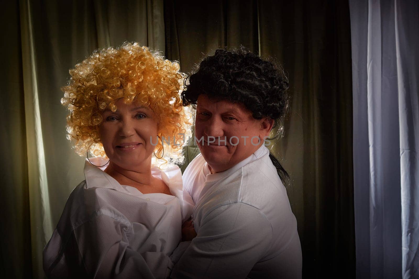 Couple enjoys a playful moment indoors, wearing vintage wigs and celebrating their love through a theatrical photoshoot in their home.