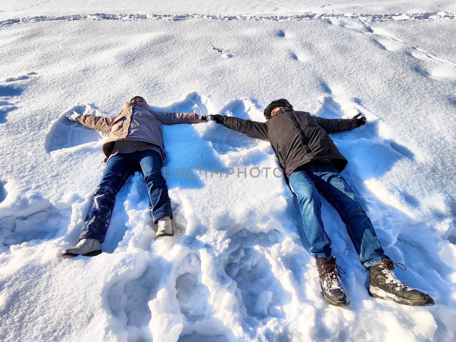 Two individuals lie on a snowy ground, creating snow angels in the bright winter sun. Their joyful expressions reflect the fun of winter activities amidst a serene snowy backdrop.