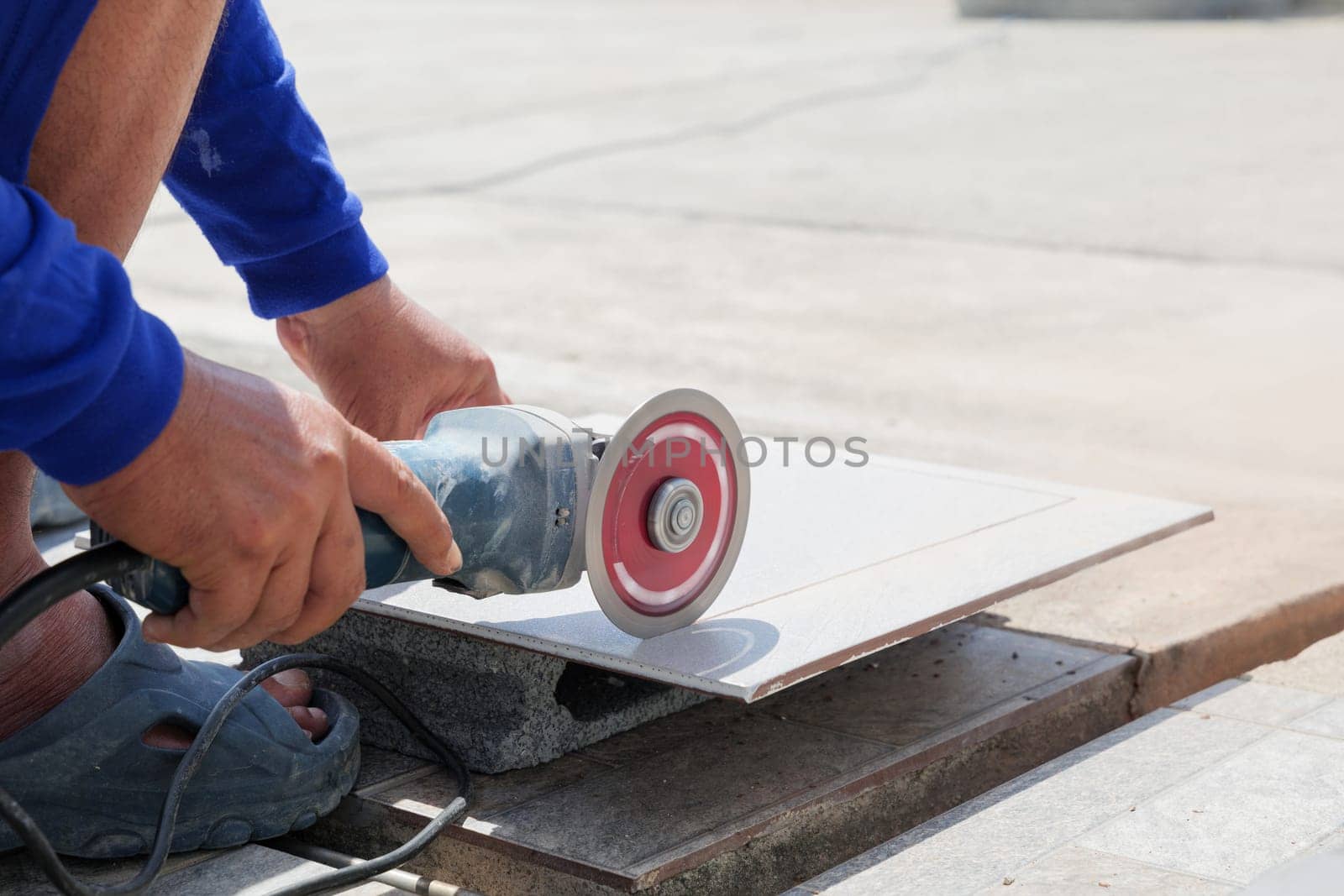 A worker wearing blue clothing operates an angle grinder to cut a gray tile on a piece of stone. The close-up view emphasizes the precision and craftsmanship of tile cutting at a construction site.